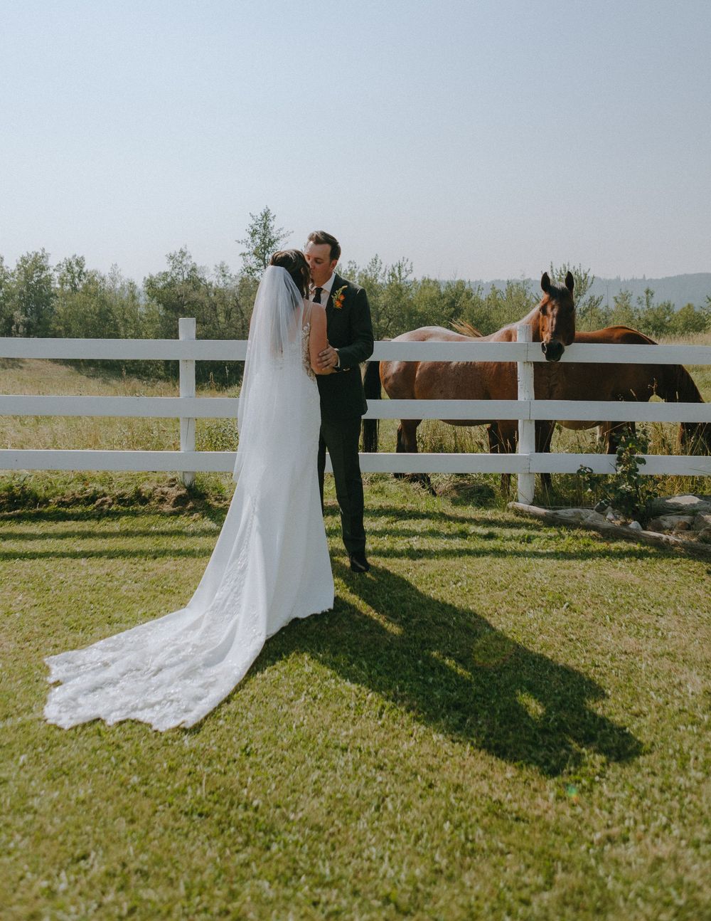 A romantic wedding moment captures a couple embracing by a white fence with a curious horse peeking over in the background.