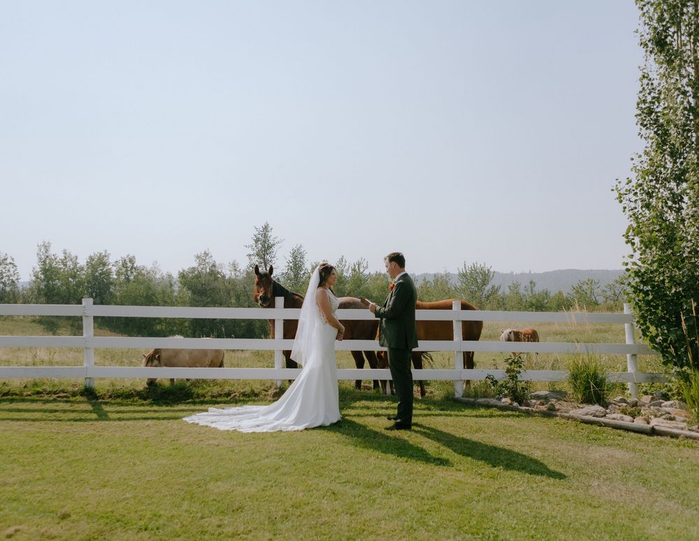 A wedding couple stands in front of a white fence on a grassy lawn with mountains visible in the distance.