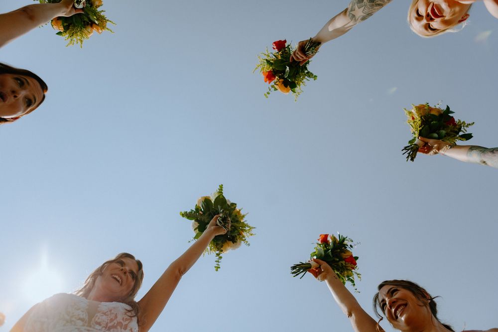 A group of bridesmaids raising colorful floral bouquets against a bright blue sky during a wedding celebration.