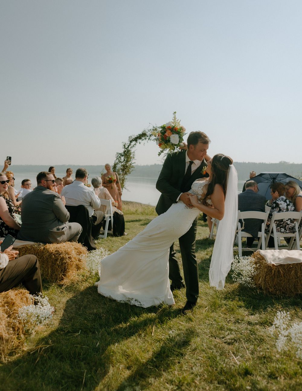 Couple sharing romantic dip kiss at sunset outdoor wedding ceremony with hay bales and floral arch by the water.