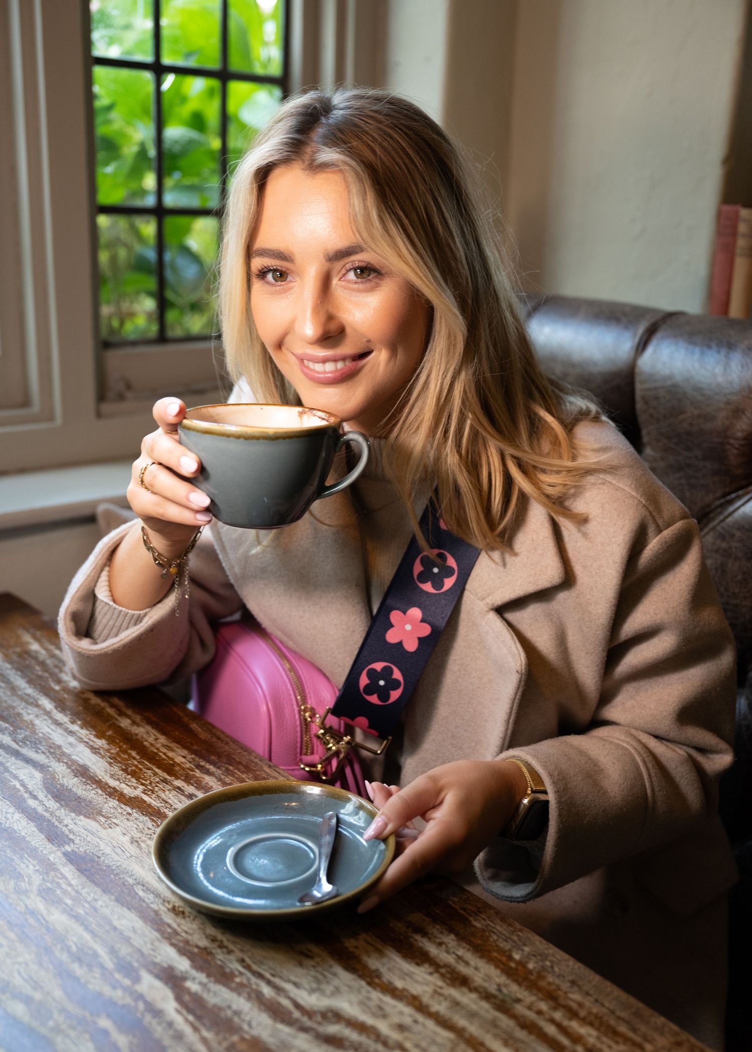 Smiling blonde woman enjoys a cappuccino at a wooden cafe table with a pink designer bag.