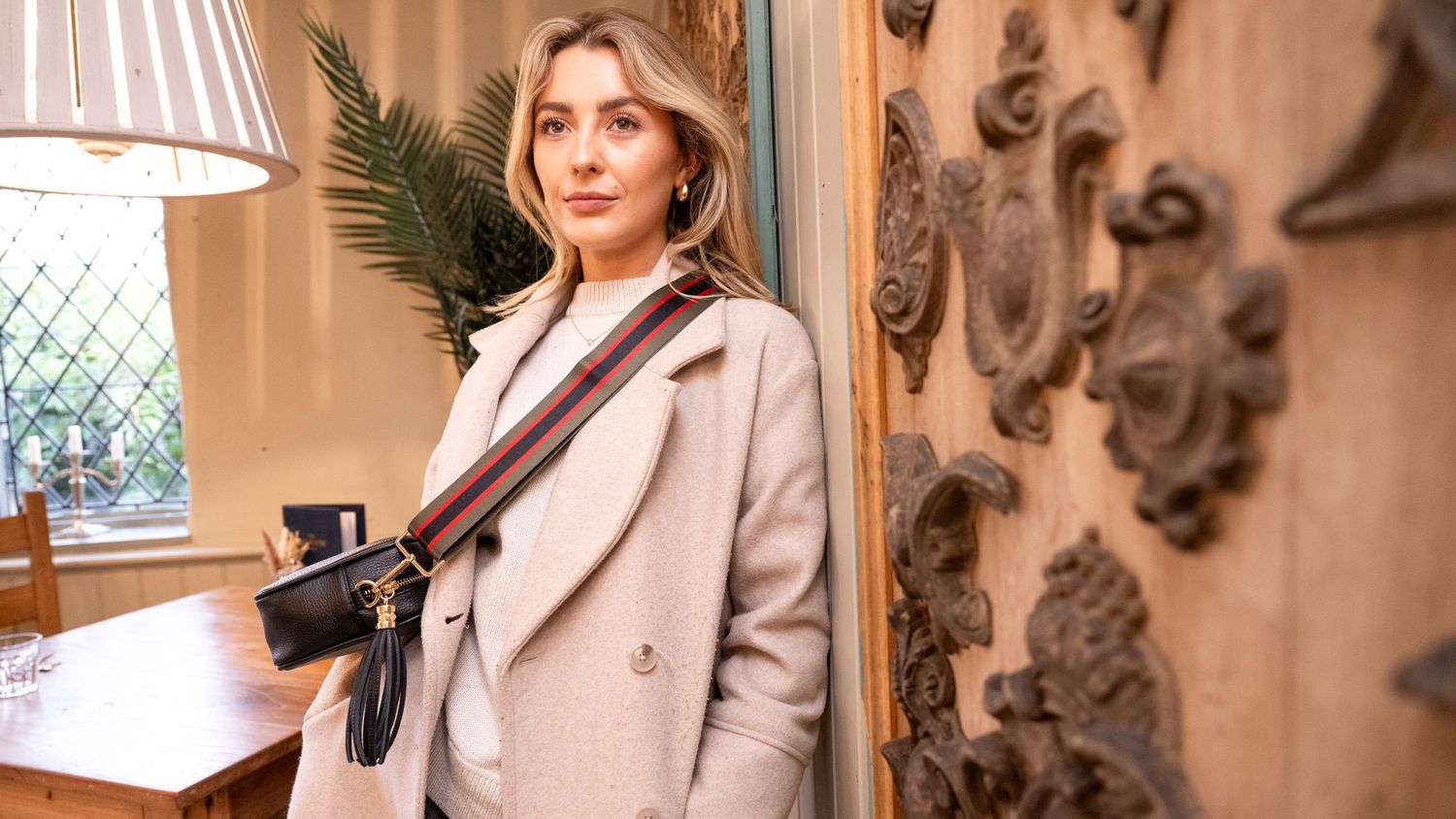Woman in beige coat carrying a small black crossbody bag with brown leather strap in elegant interior.