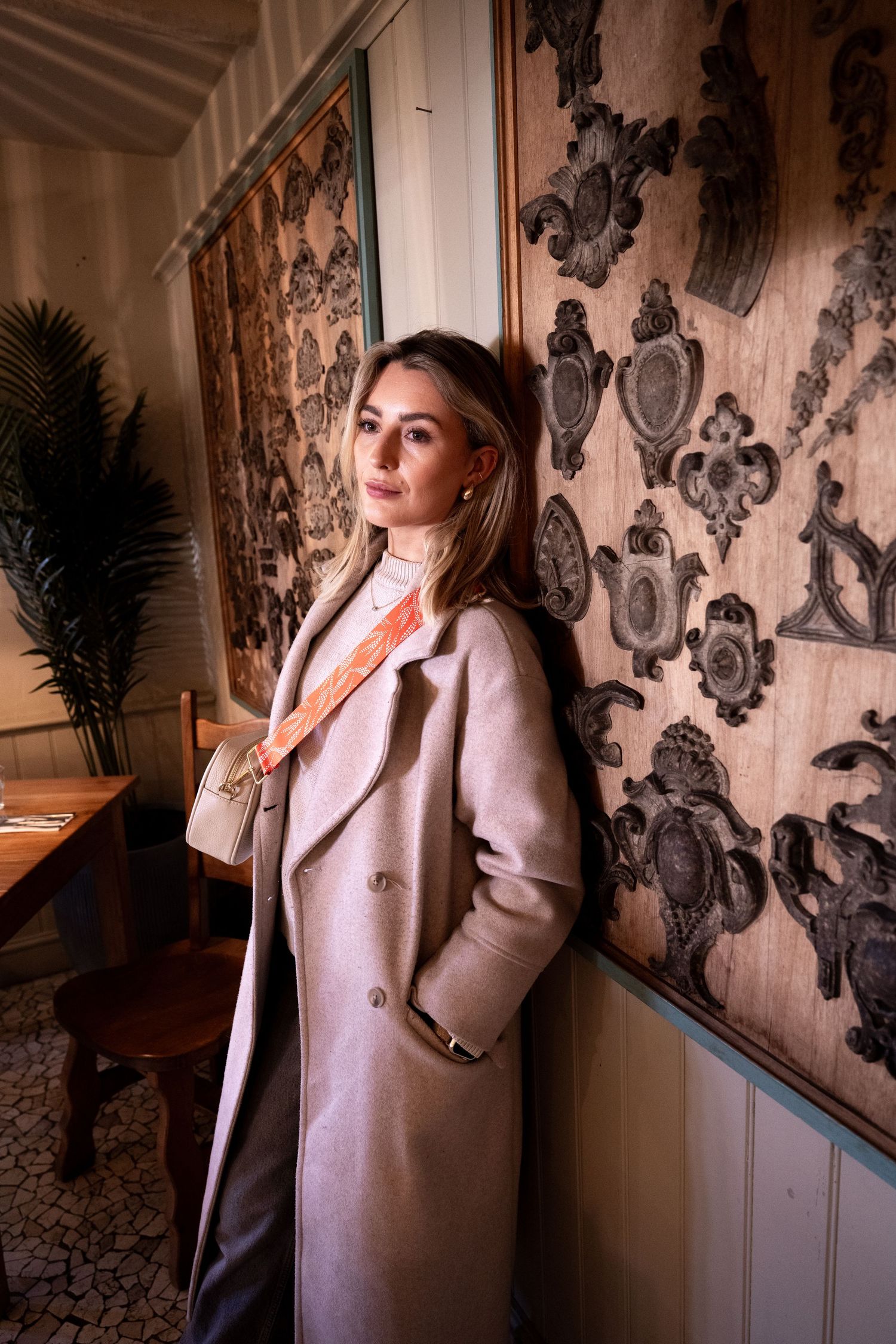 Elegant woman in beige wool coat with orange silk neck scarf leaning against ornate wall.
