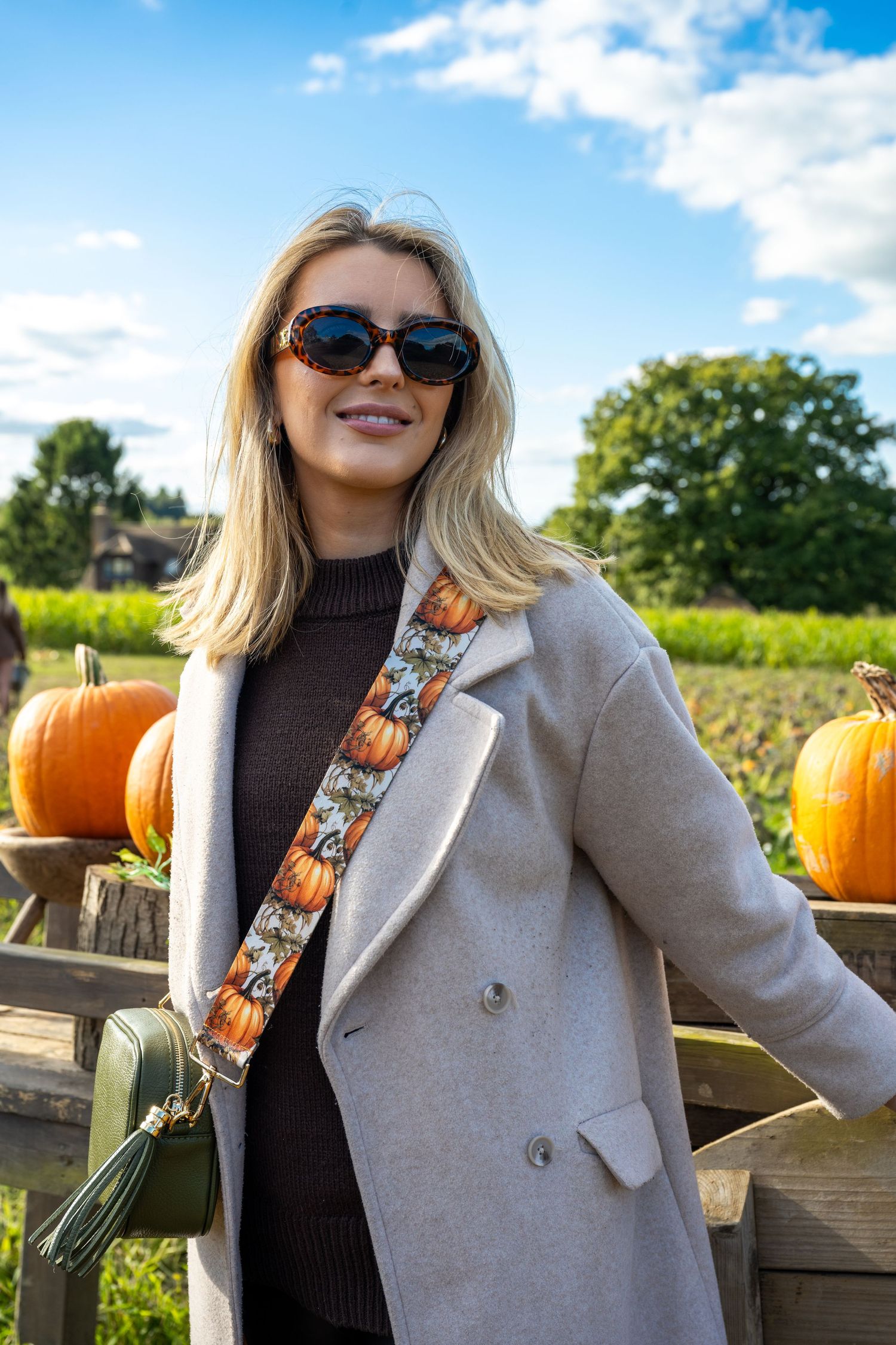 Smiling blonde woman in beige coat with pumpkin print bag strap at autumn pumpkin patch.