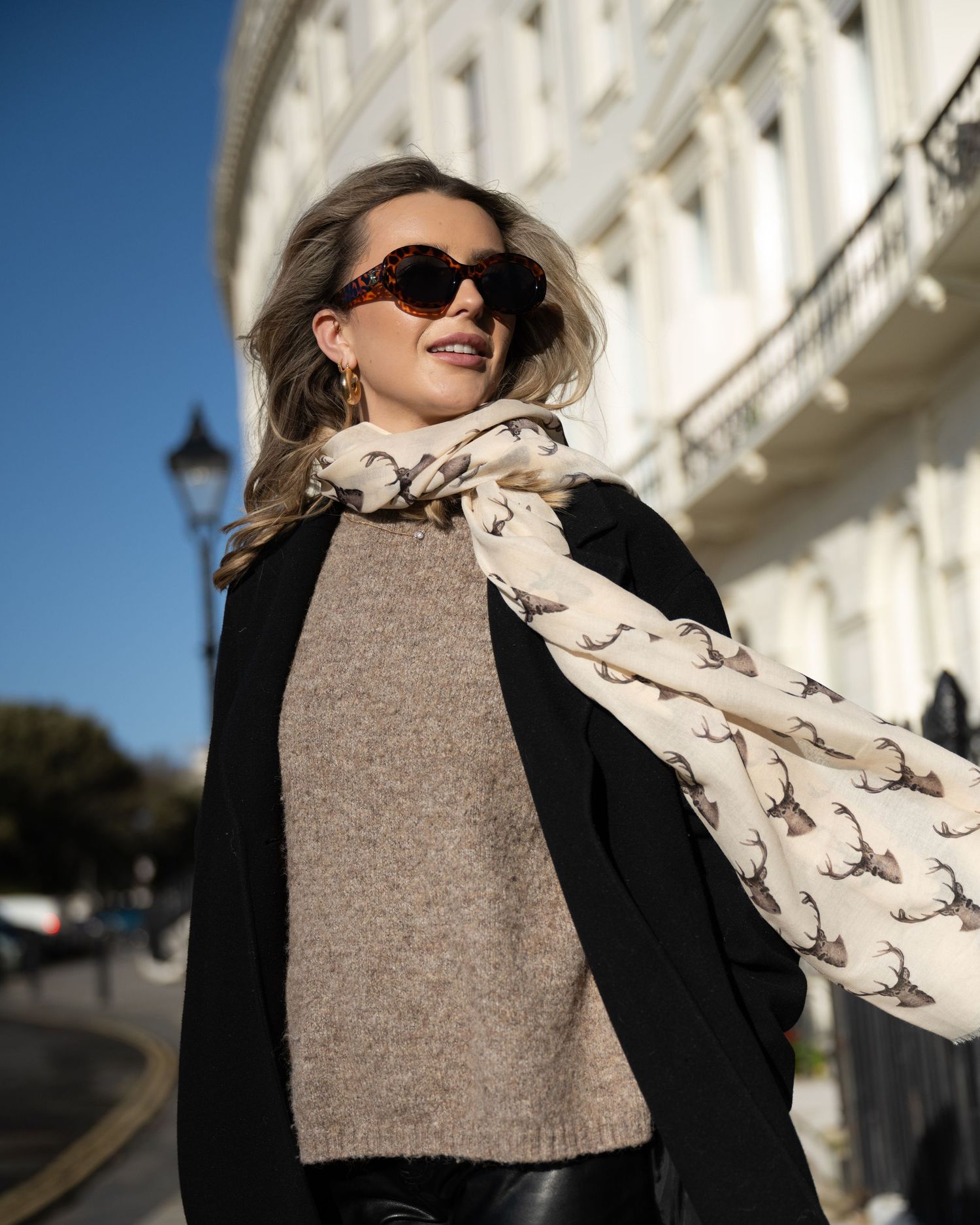 Close-up of woman in black coat and beige sweater wearing a bird-print scarf and cat-eye sunglasses outdoors.