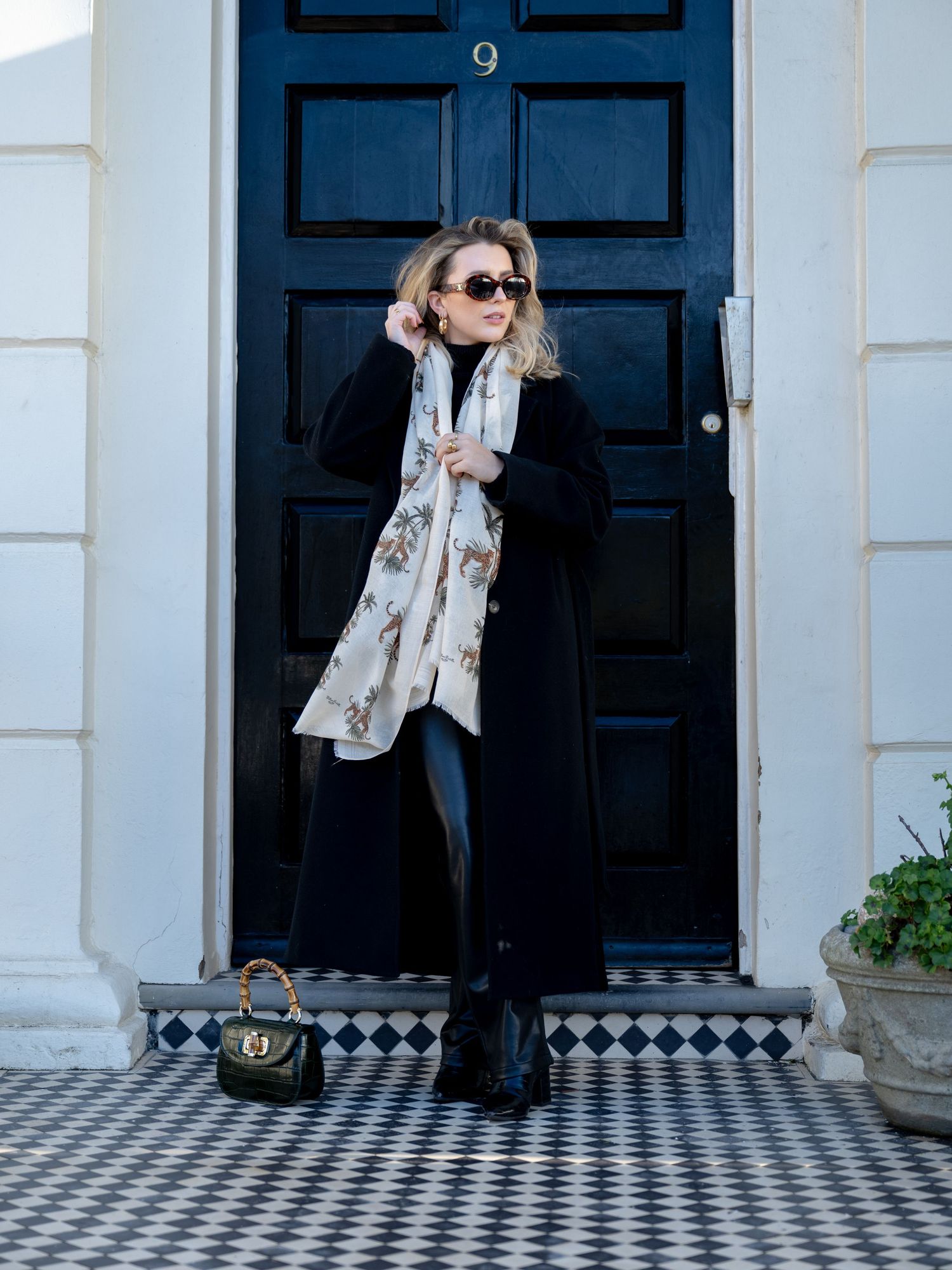 Woman in black coat with floral scarf and sunglasses standing before a dark blue door on a tiled doorstep.
