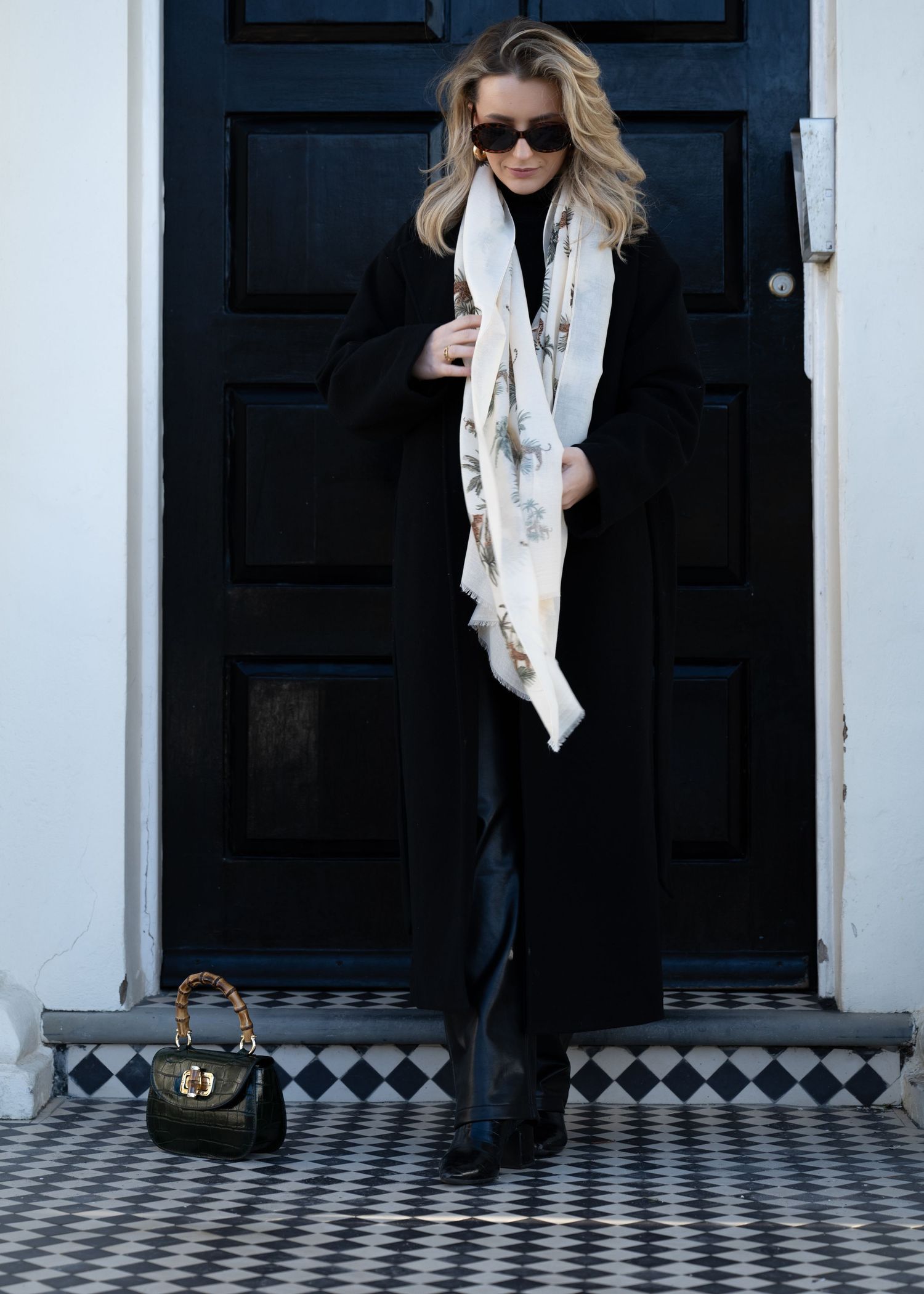 Blonde woman in long black coat with a light floral scarf and small handbag standing at a black door.