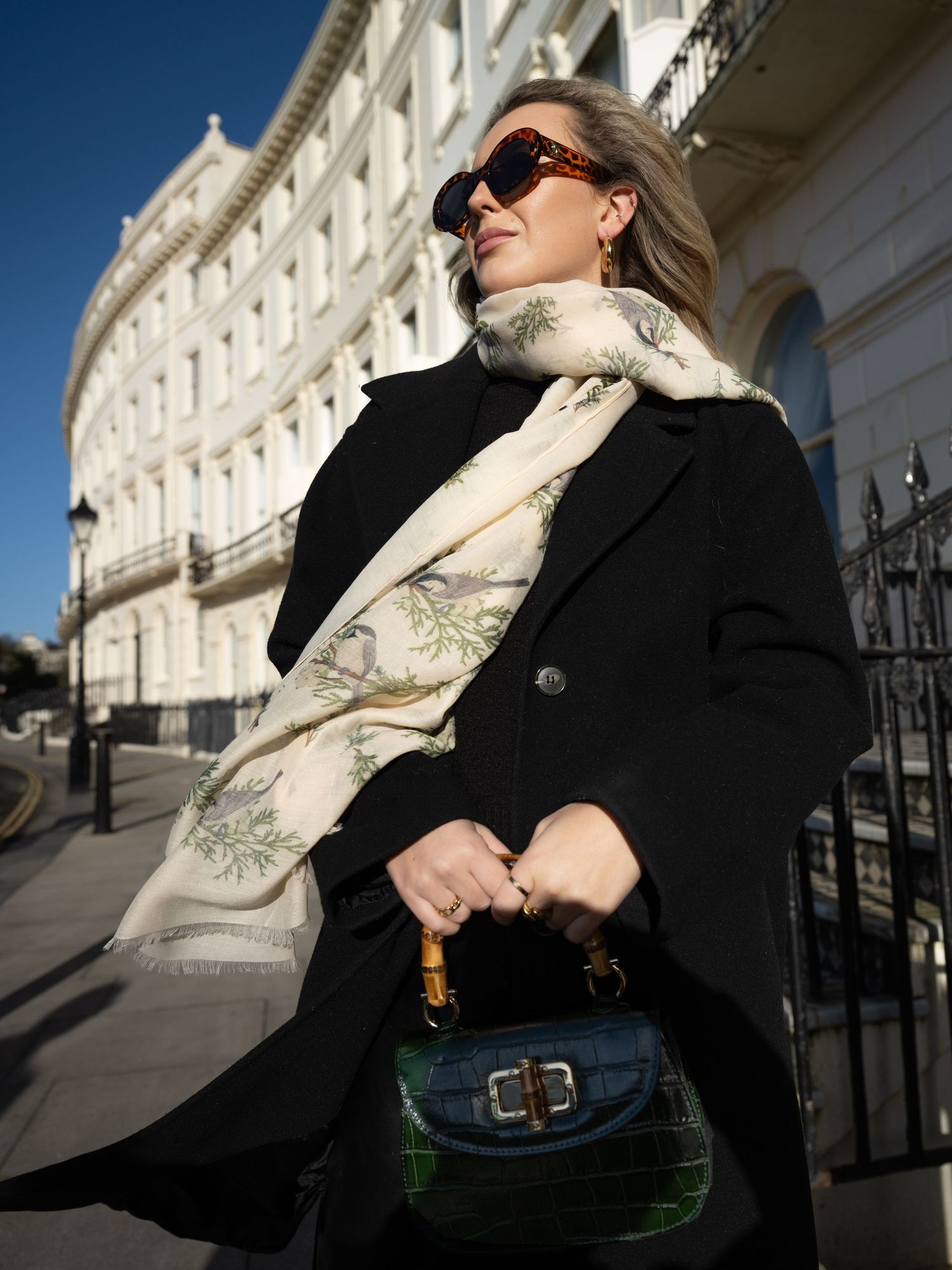 Blonde woman in black coat wearing a botanical print scarf and tortoiseshell sunglasses on a sunny street.