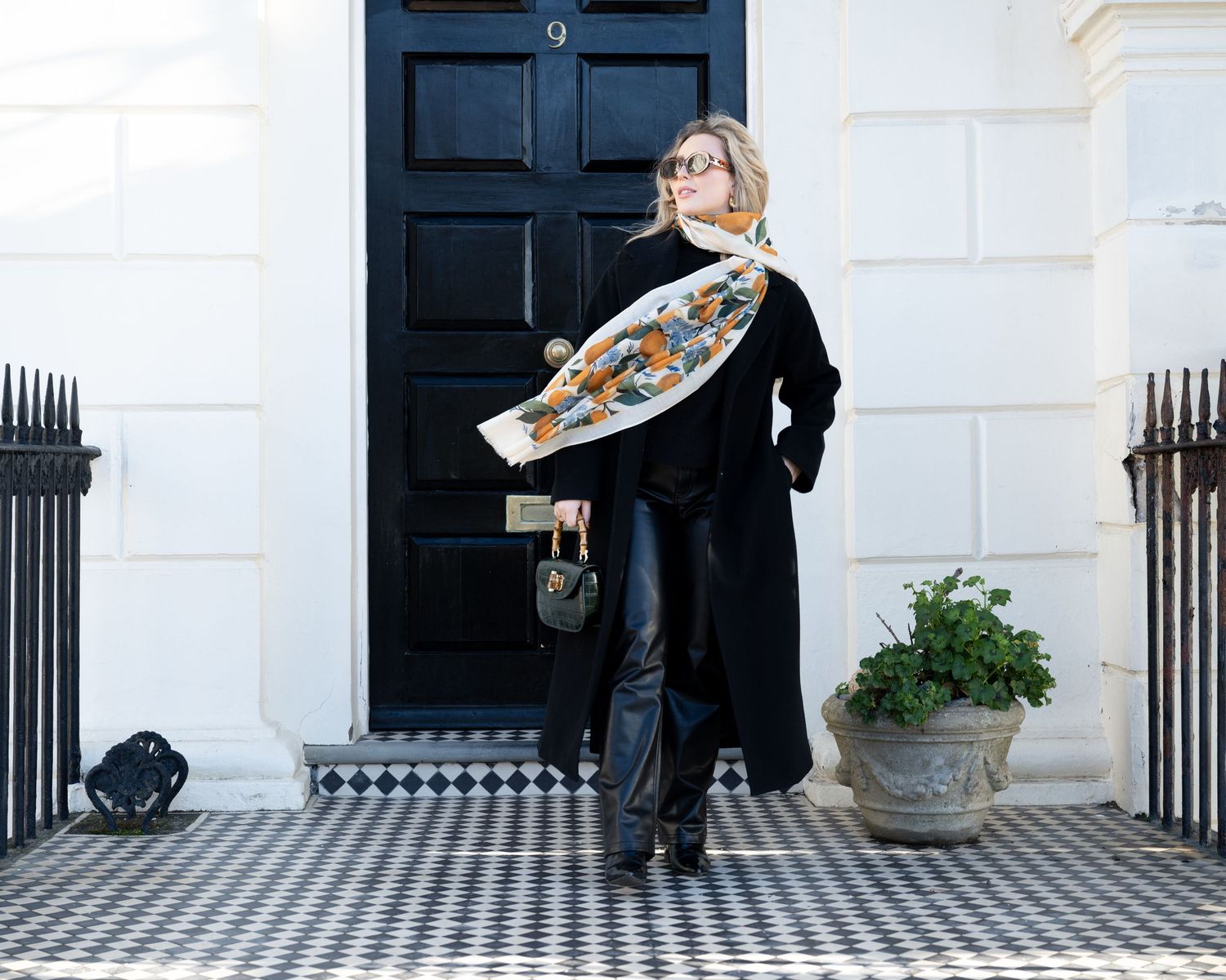 Woman in black coat holding an orange floral scarf open while standing on a black and white tiled doorstep.