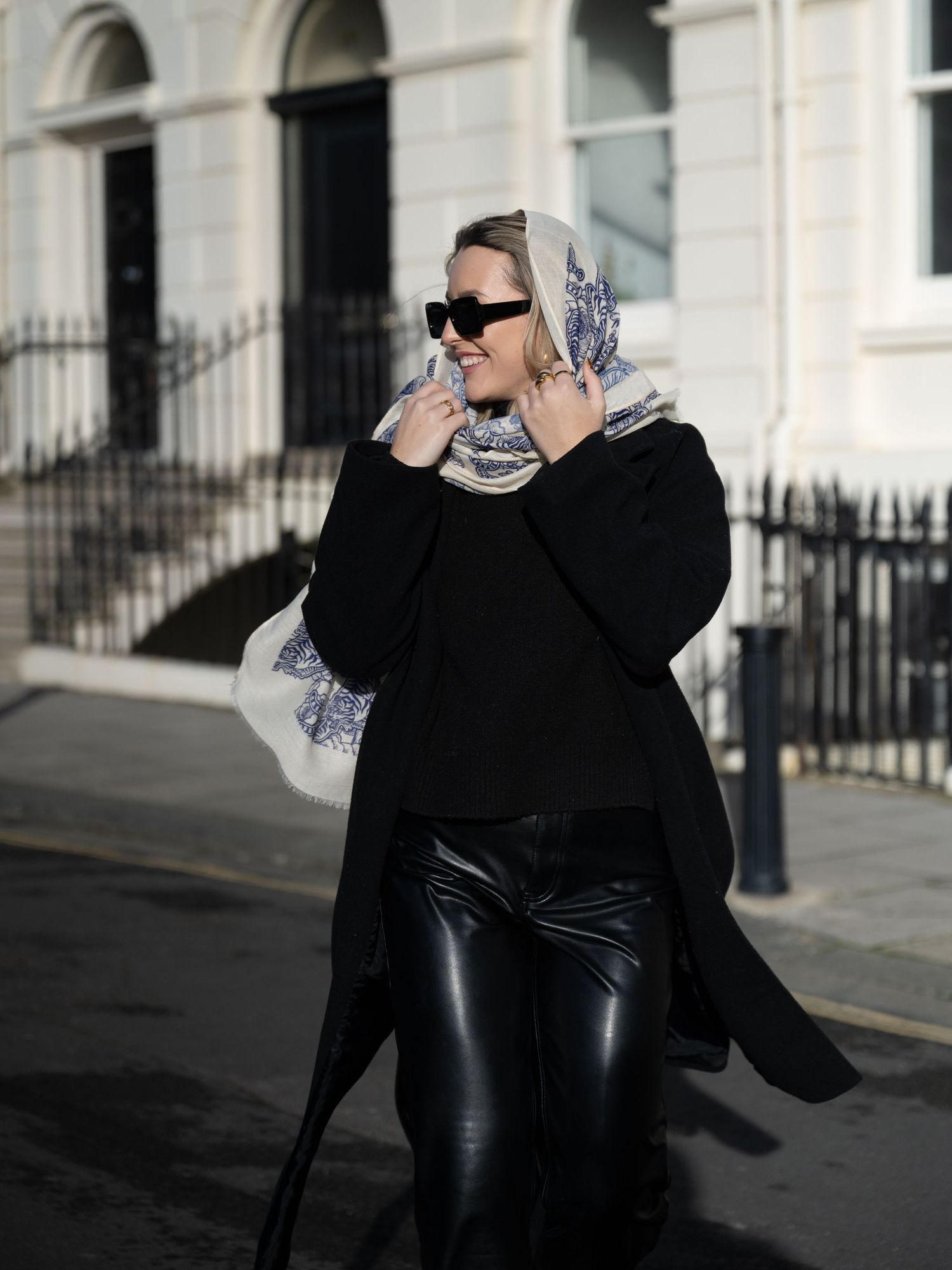 Smiling woman in black outfit with a printed scarf tied as headscarf walking on a bright urban street.