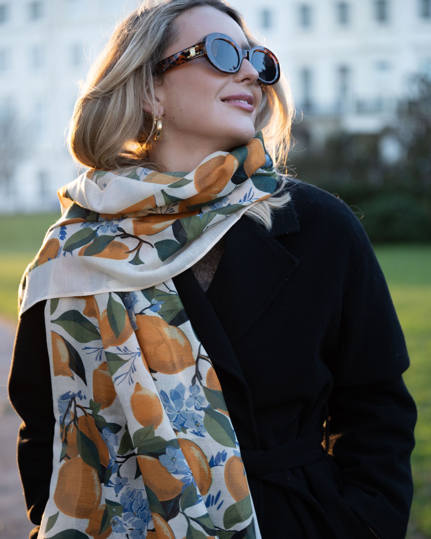 Woman in black coat wearing an orange and blue floral print scarf with round sunglasses in a park setting.