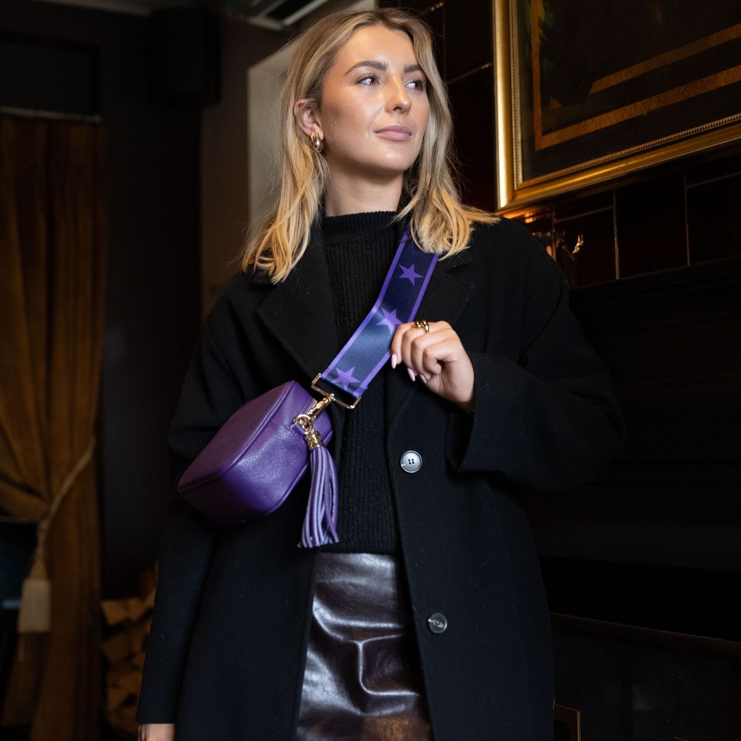 Blonde woman in black coat with purple leather crossbody tassel bag on wide fabric strap.