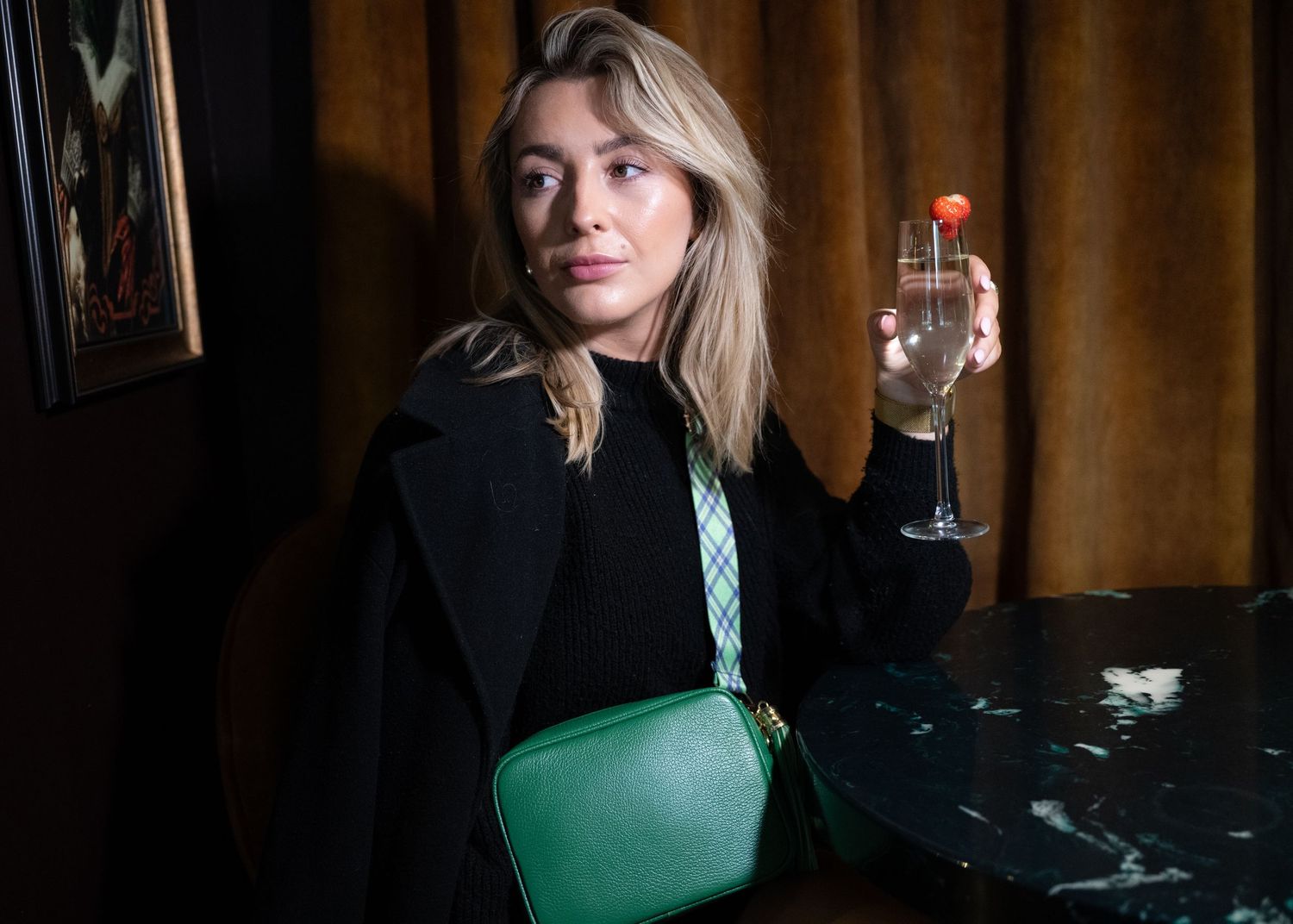 Woman in black coat holding cocktail with green leather crossbody bag on plaid strap at bar.