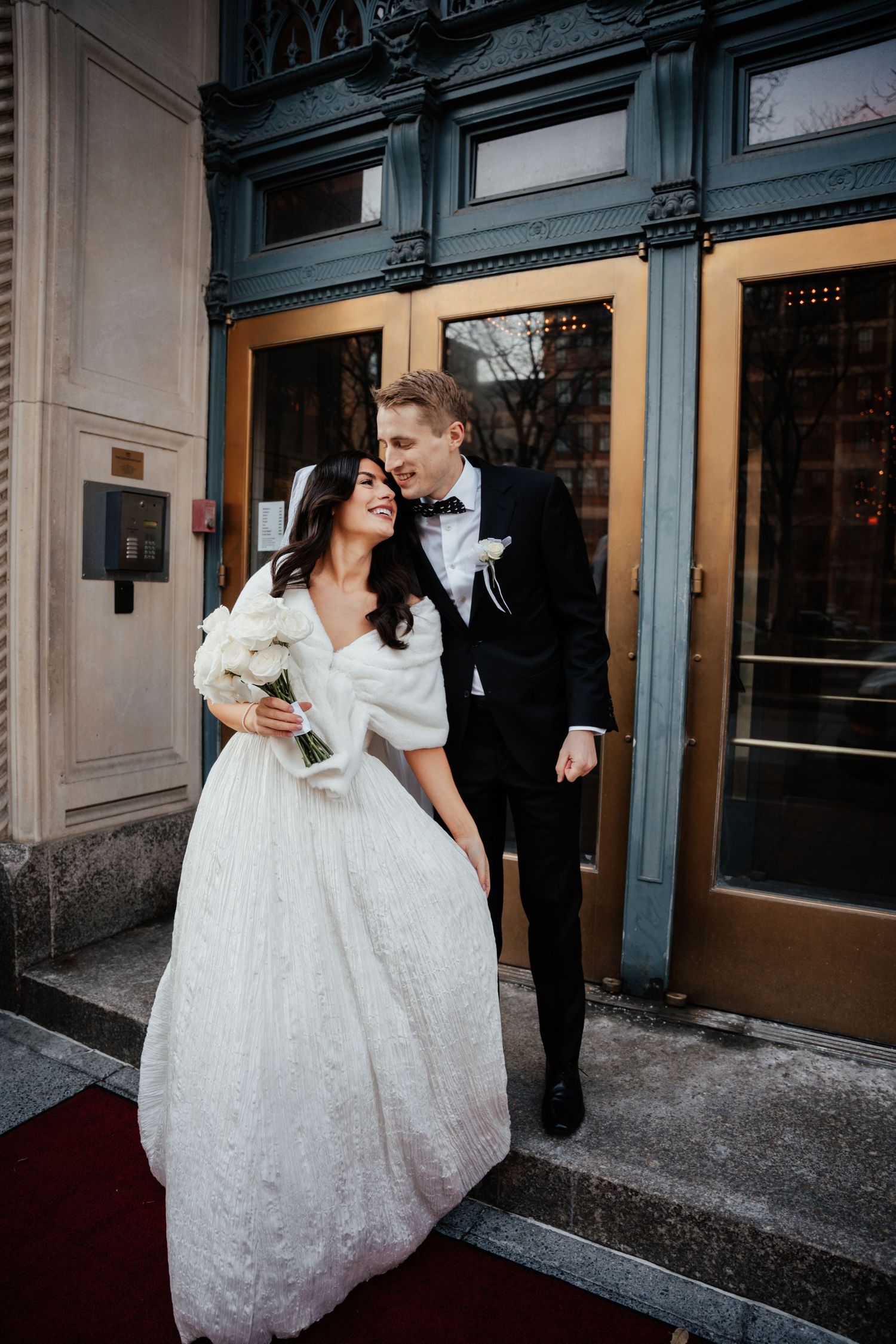 A couple in wedding attire stands in front of a historic building entrance with blue-painted details and ornate architecture.