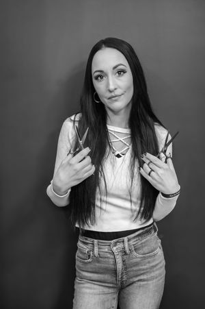 Hair stylist in white top and jeans makes rock gesture while posing against dark background.