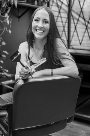 Hairstylist smiles while holding styling tools in black and white portrait.