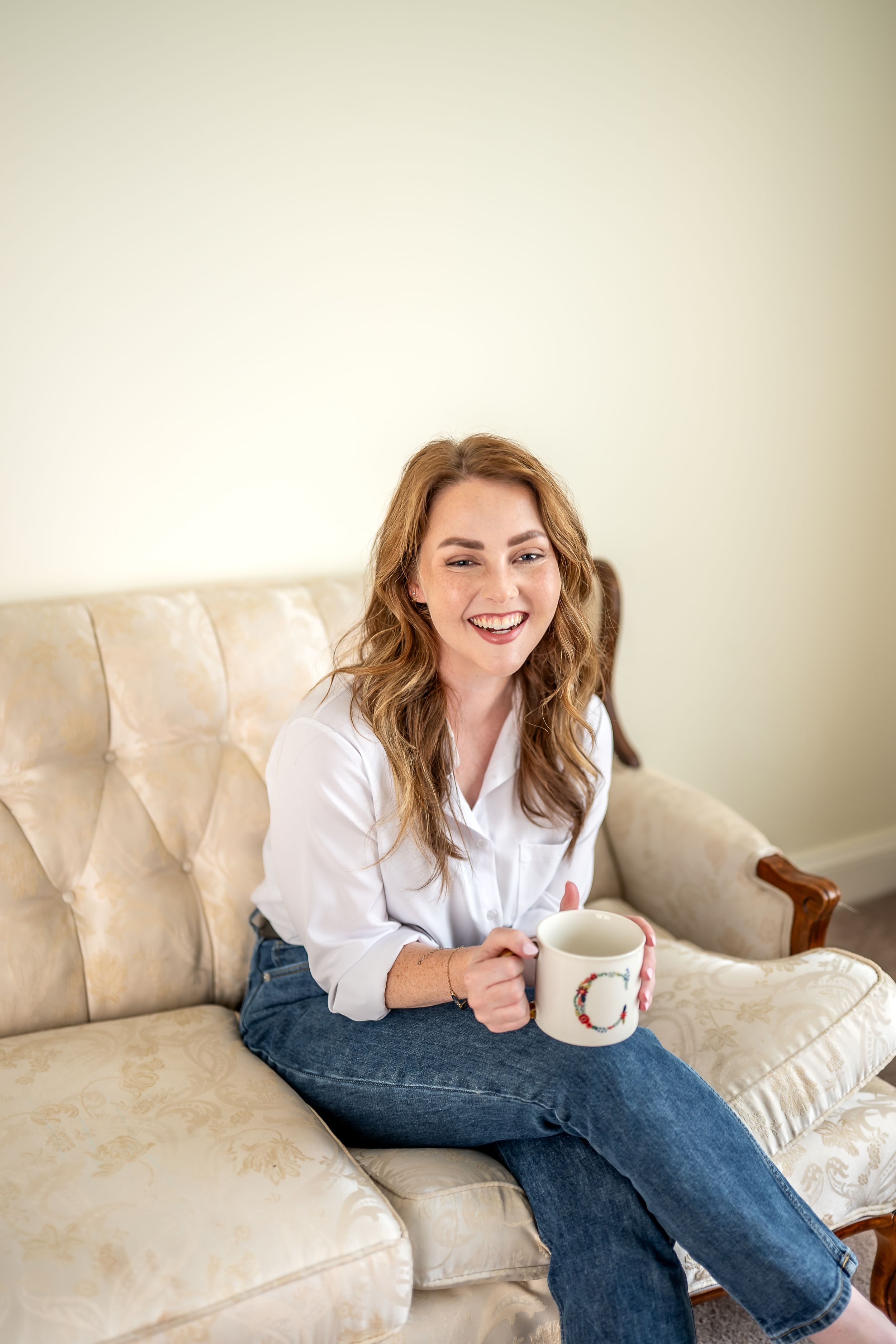 Person in white shirt and blue jeans relaxing on light-colored couch with coffee mug.