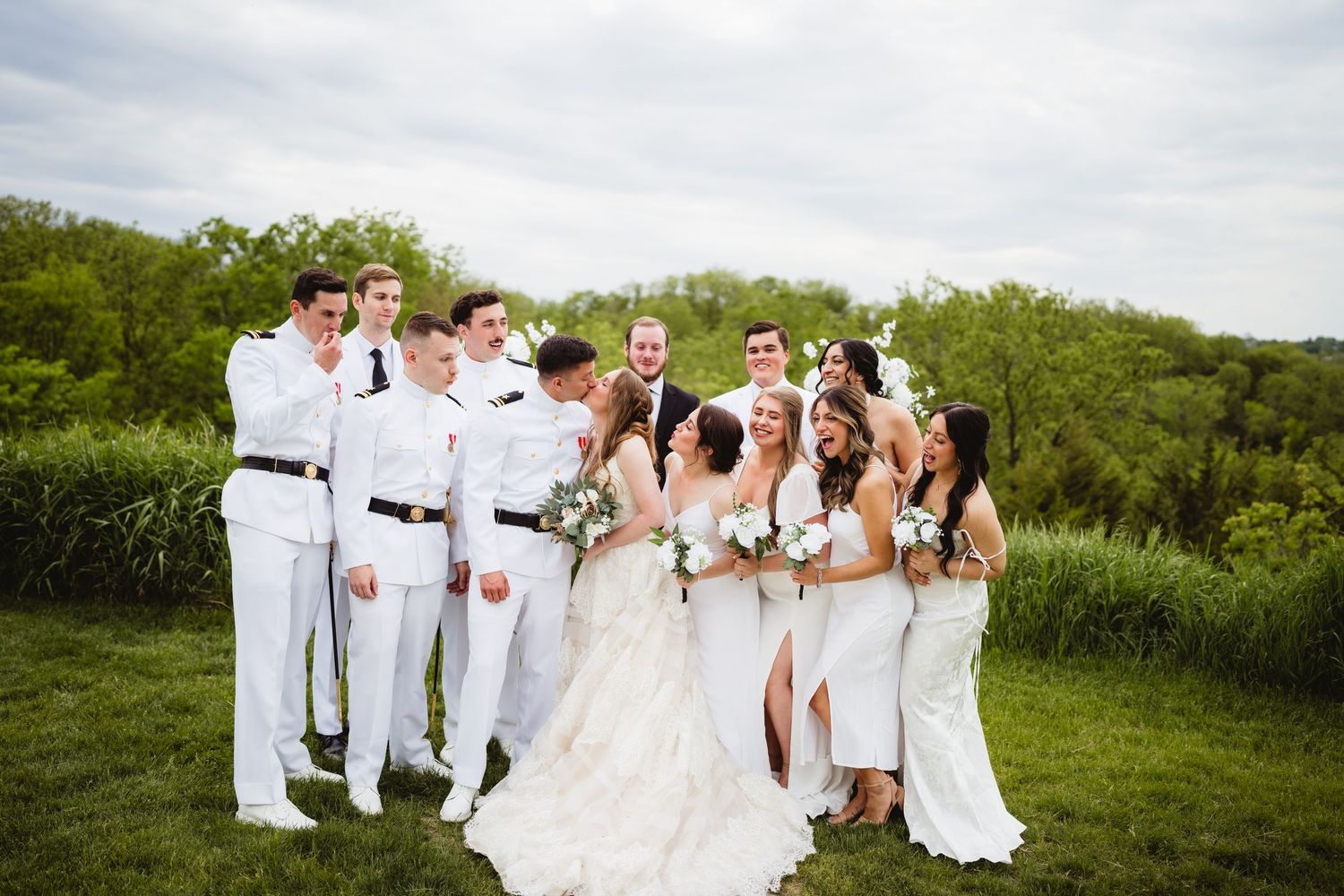 A wedding party in all white attire poses together on a grassy hill against a backdrop of green trees.