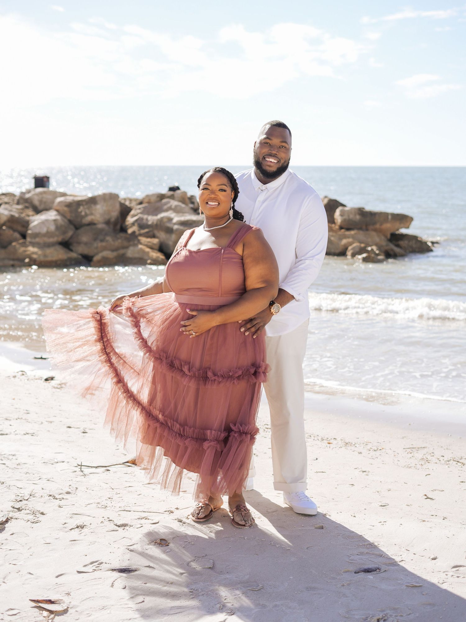 A couple stands together on a beach with rocky shoreline, the woman wearing a flowing pink dress and man in white attire.