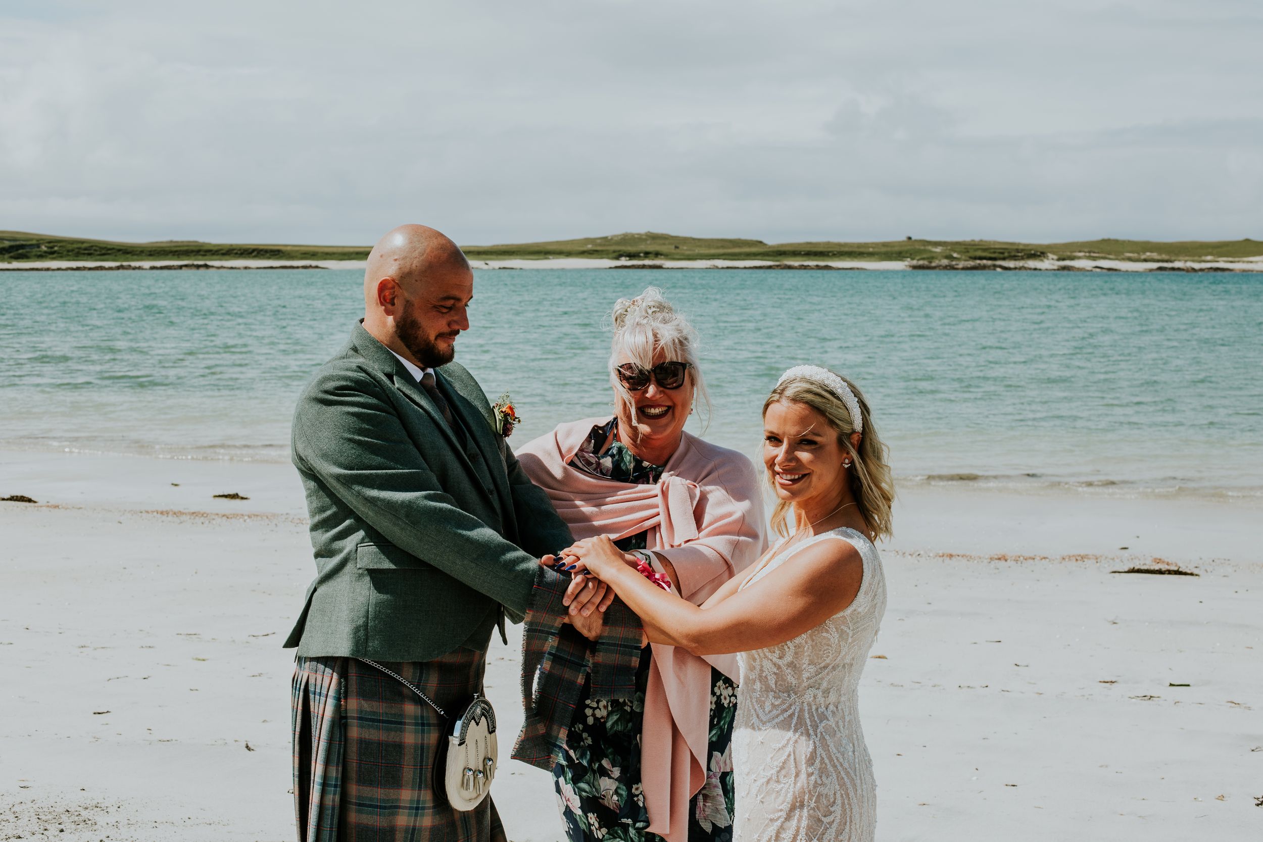 Three people stand together in a ceremony on a scenic beach with turquoise water in the background.