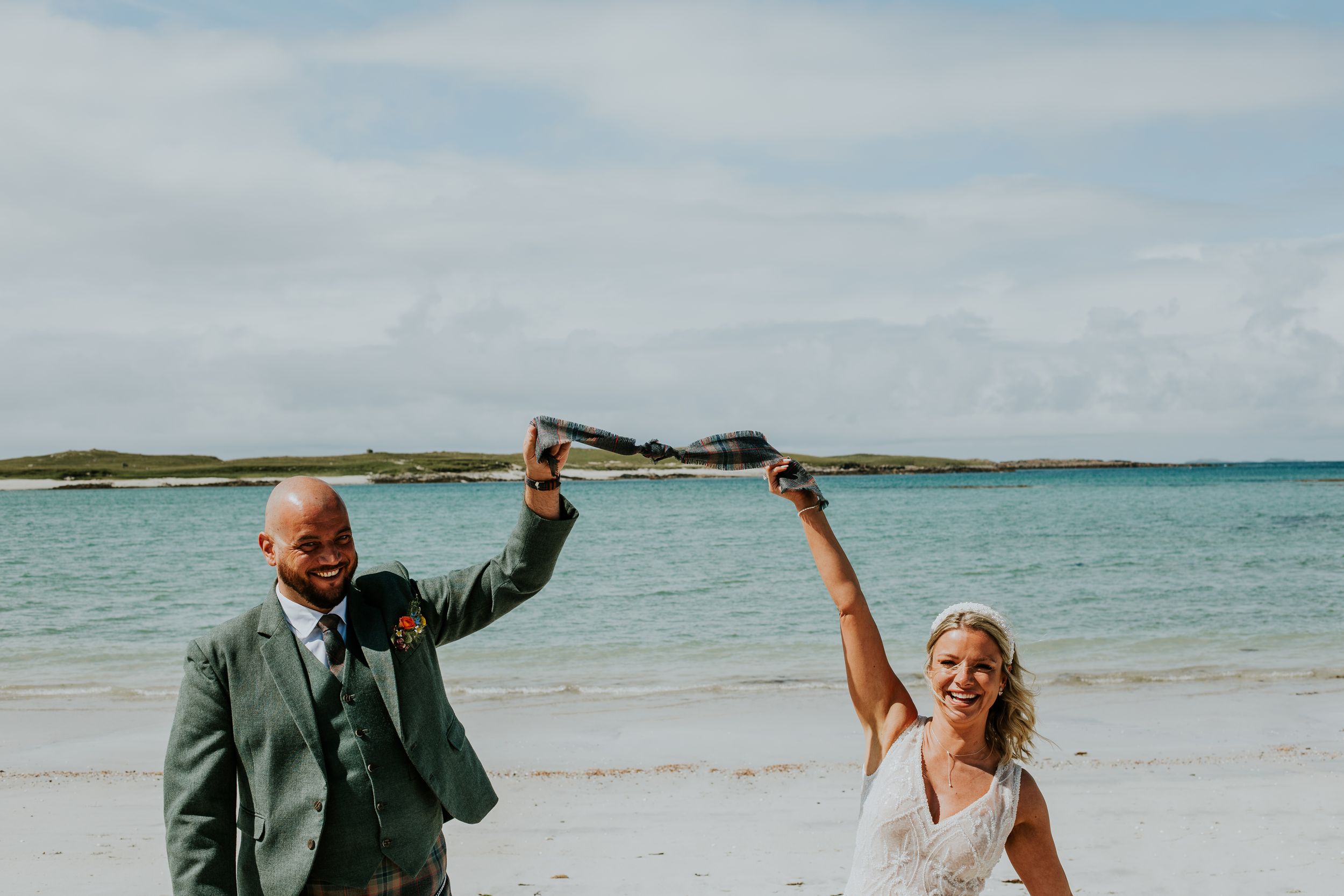 Couple celebrating on sandy beach with joyful dance moves in wedding attire.
