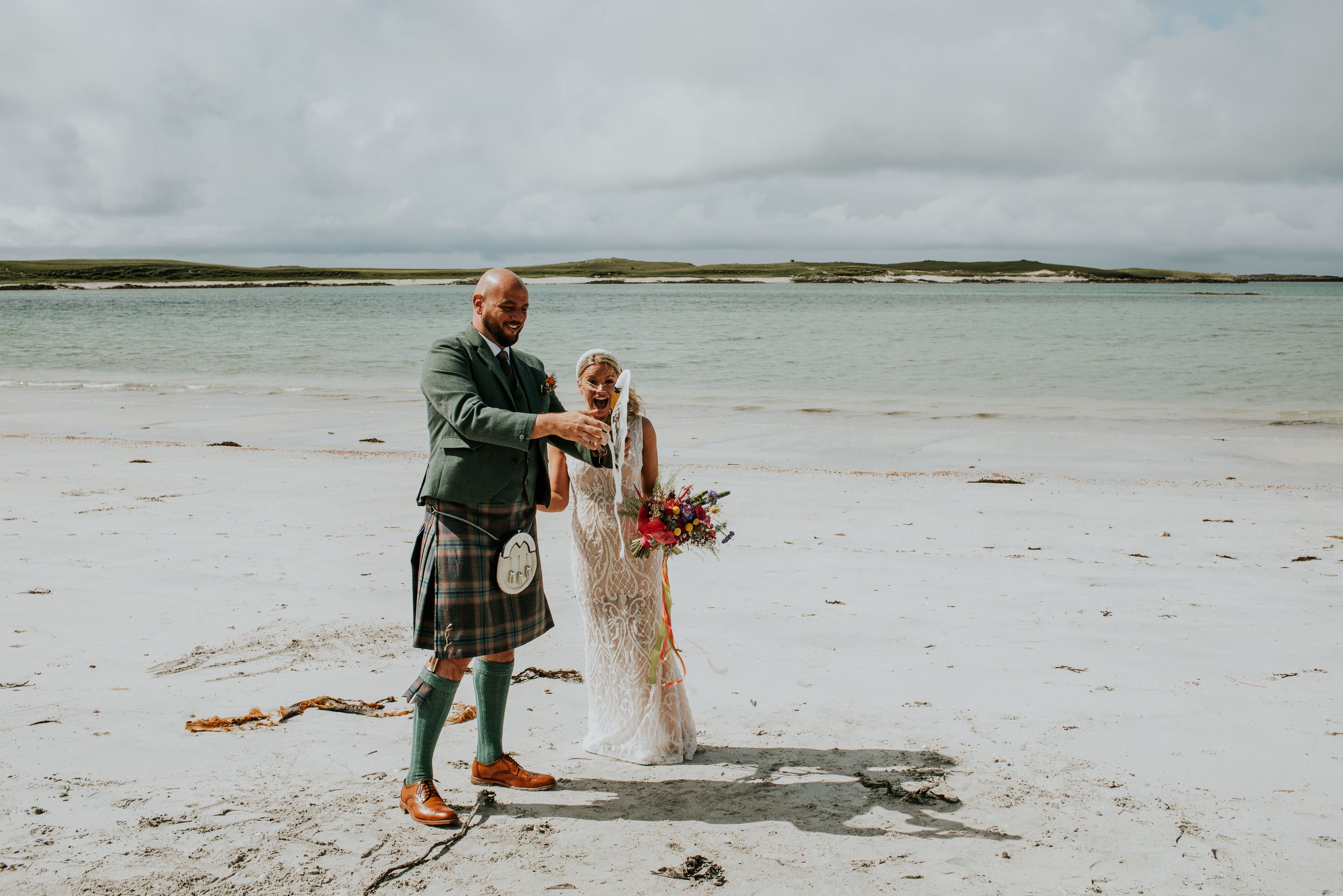 Newlyweds walking together on a pristine white sand beach with traditional Scottish wedding attire.