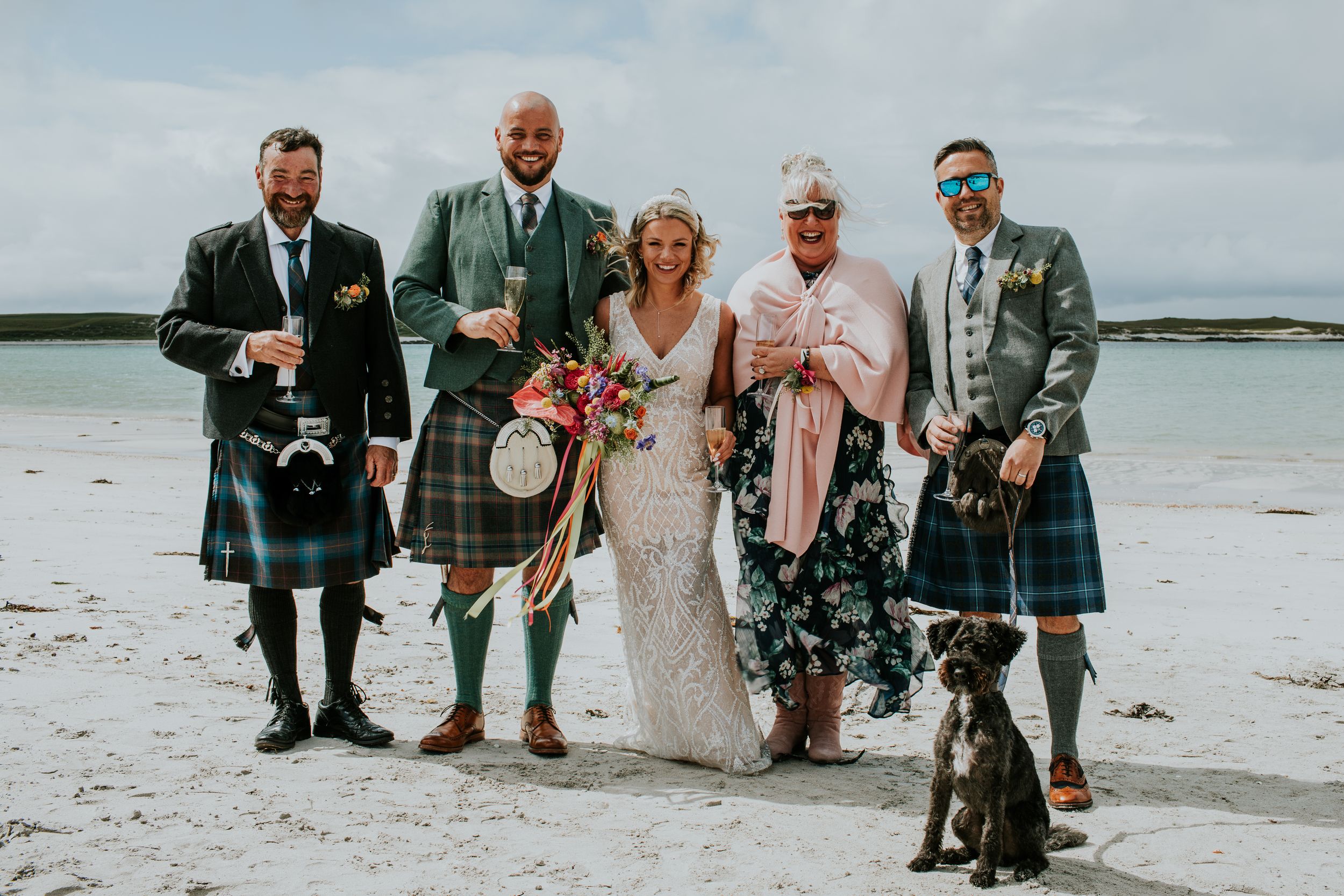 Wedding party wearing traditional Scottish kilts posing on beach with dog.