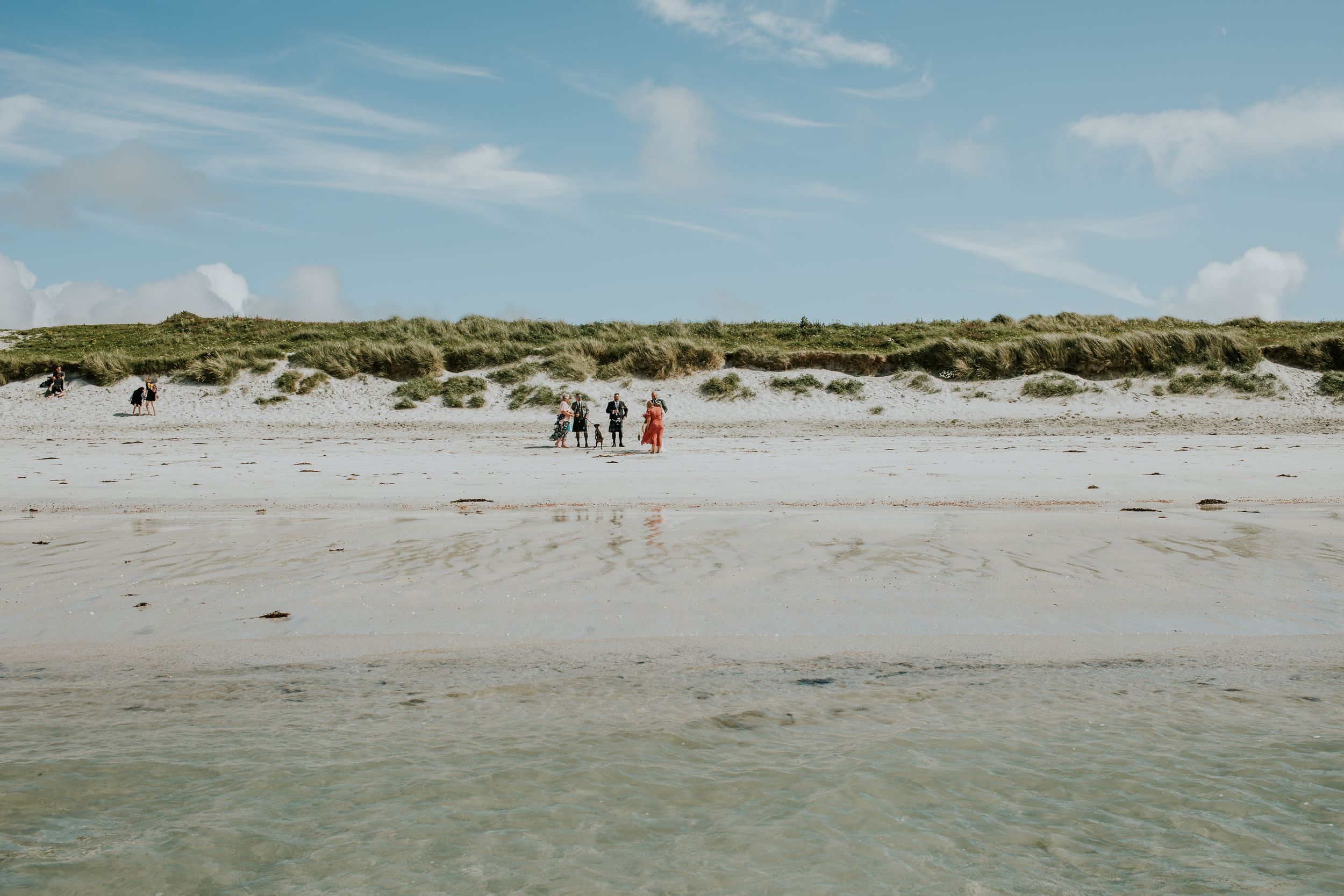 Wide shot of people walking on a remote beach with sand dunes and blue water.