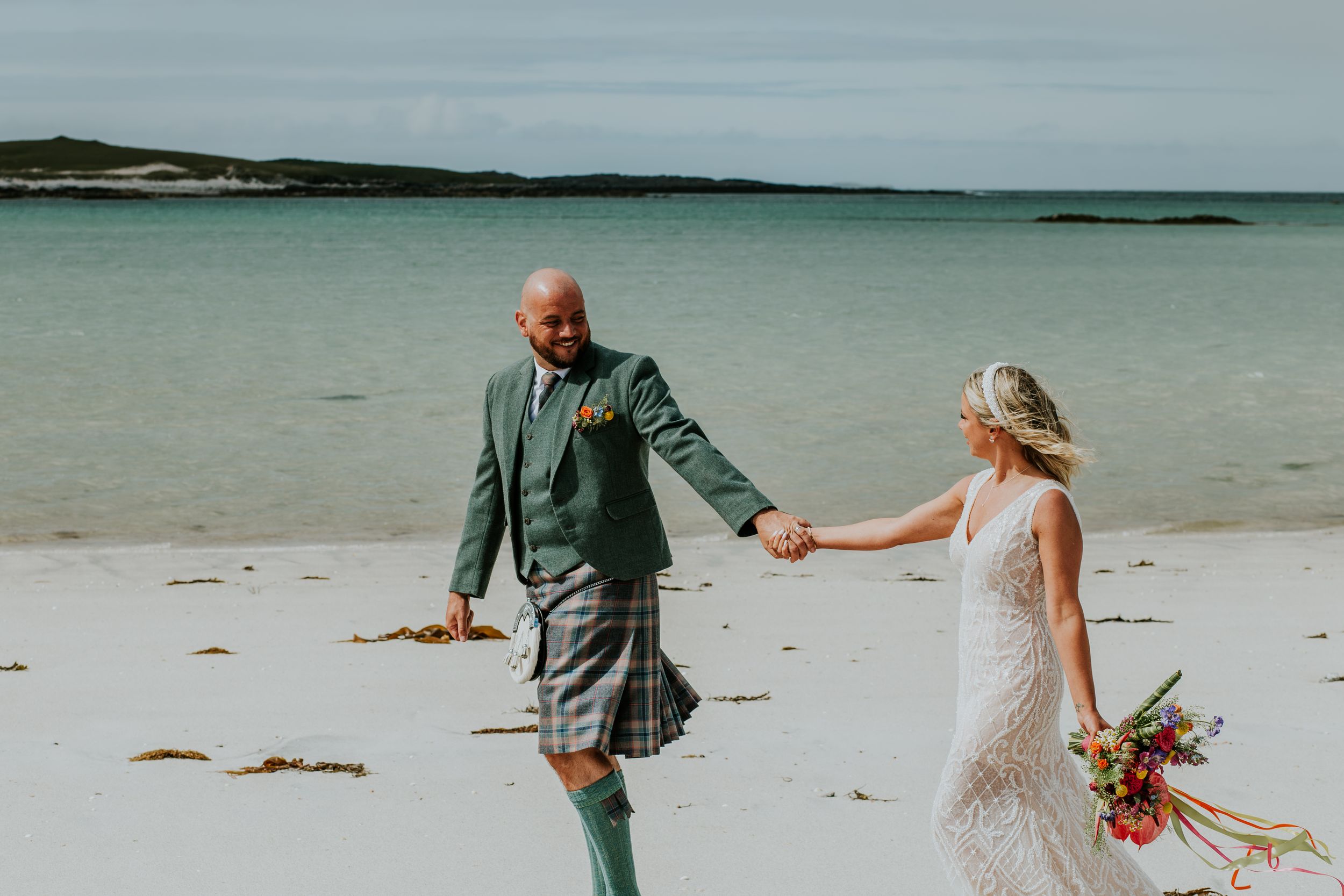 A couple walks hand in hand on a beach during their Scottish-themed wedding celebration.