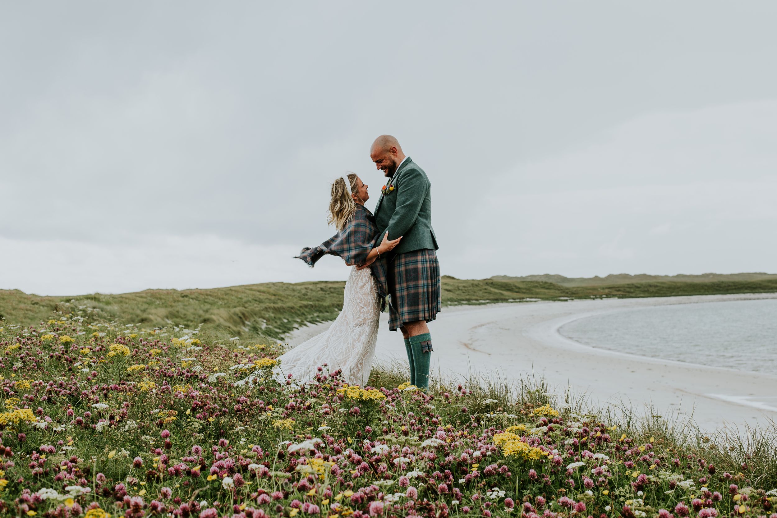 A couple shares an intimate moment on a grassy hillside overlooking a coastal beach in Scotland.