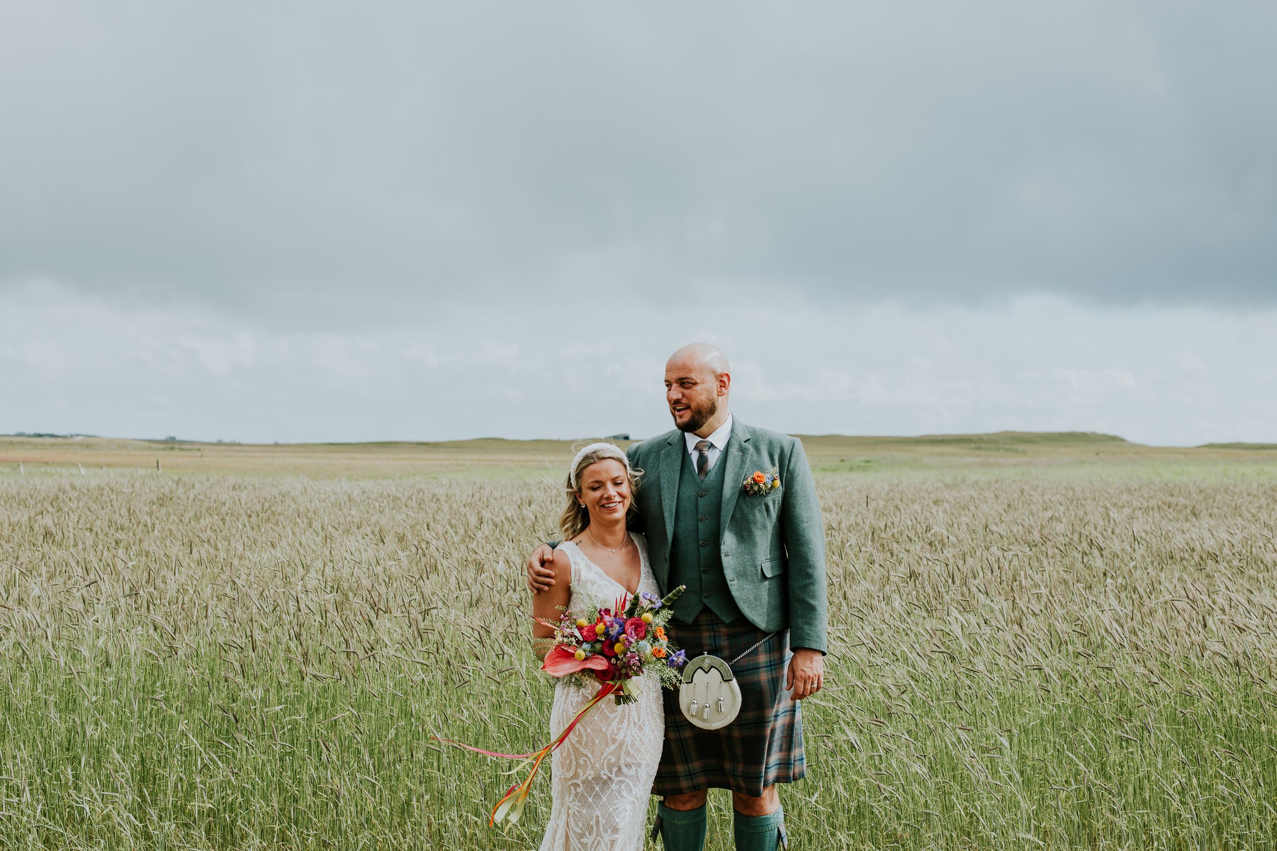 Wedding couple poses in a scenic field wearing traditional Scottish formal wear with a colorful bouquet.