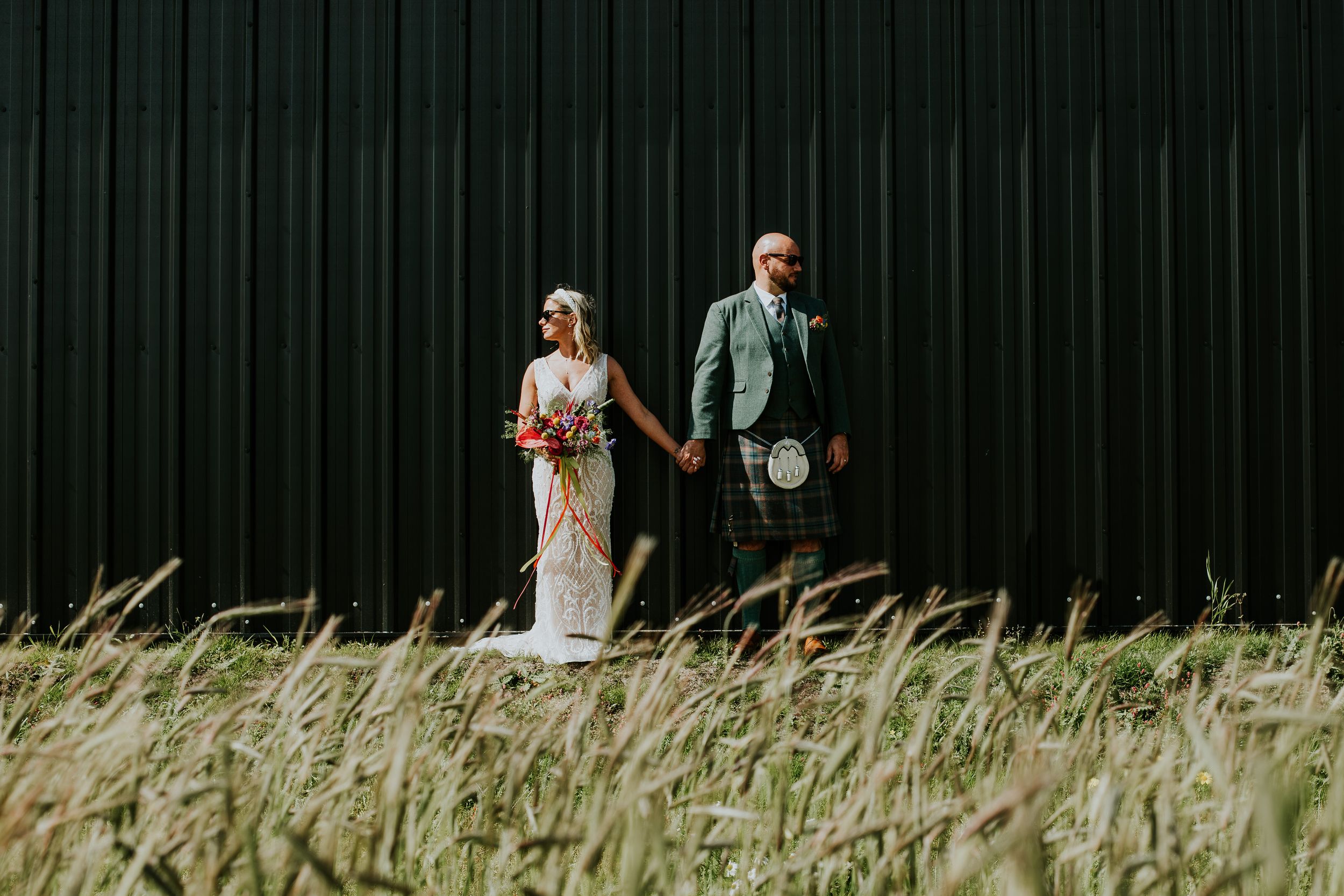 Artistic wedding portrait against black metal wall with tall grass in foreground.