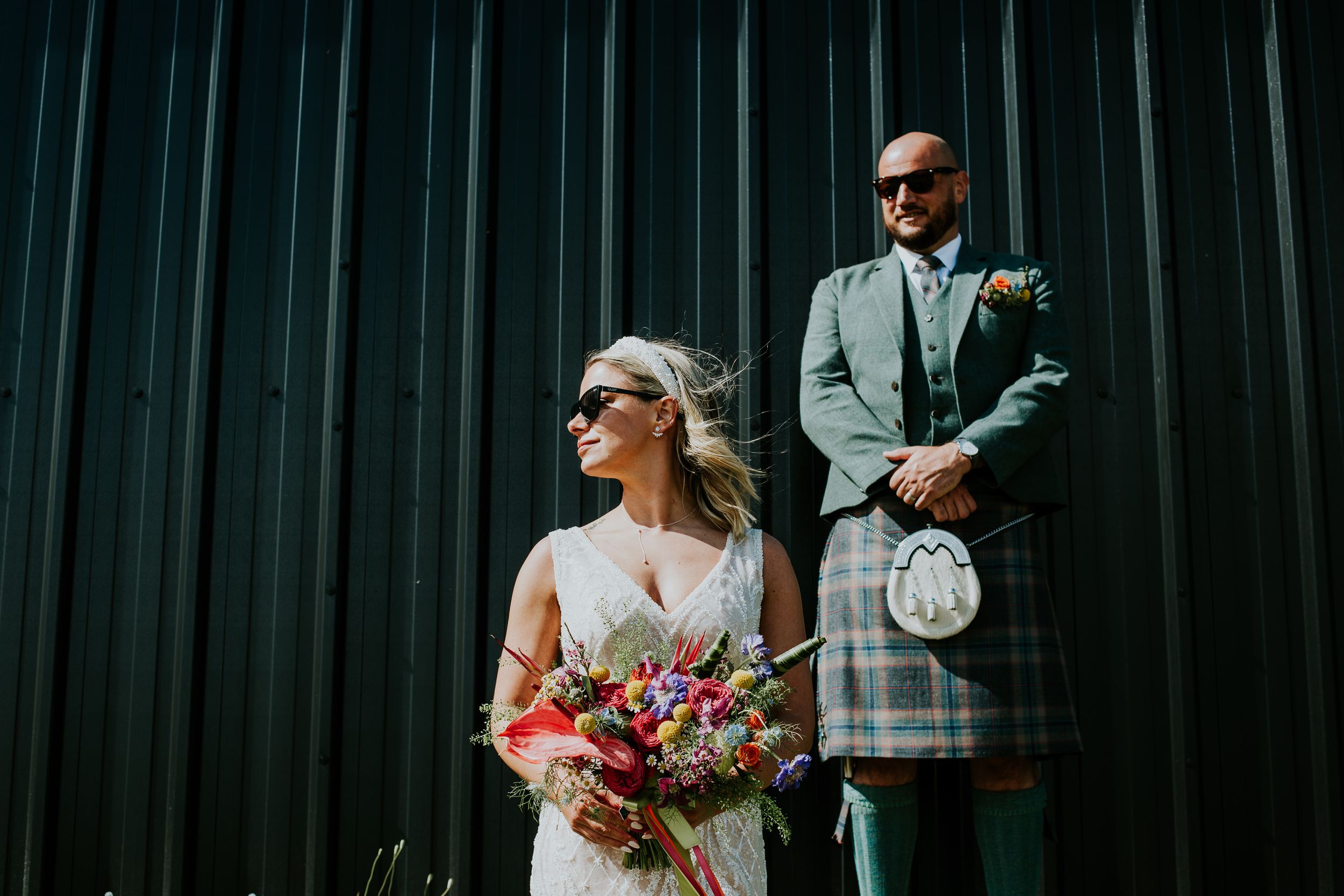Stylish wedding couple in sunglasses pose against dark metal wall wearing Scottish formal wear.