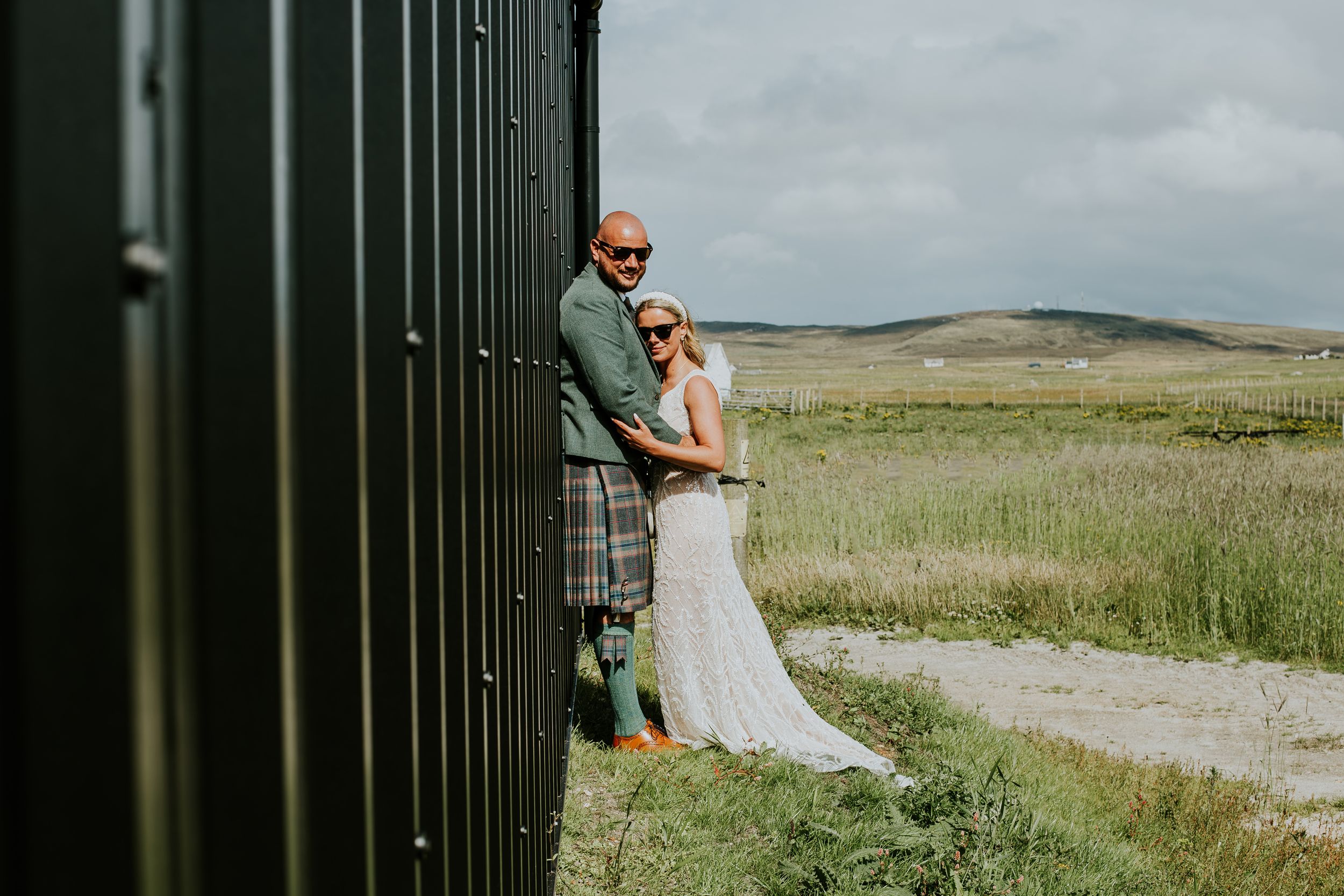 A wedding couple poses playfully against a dark wall with rolling hills in the background.