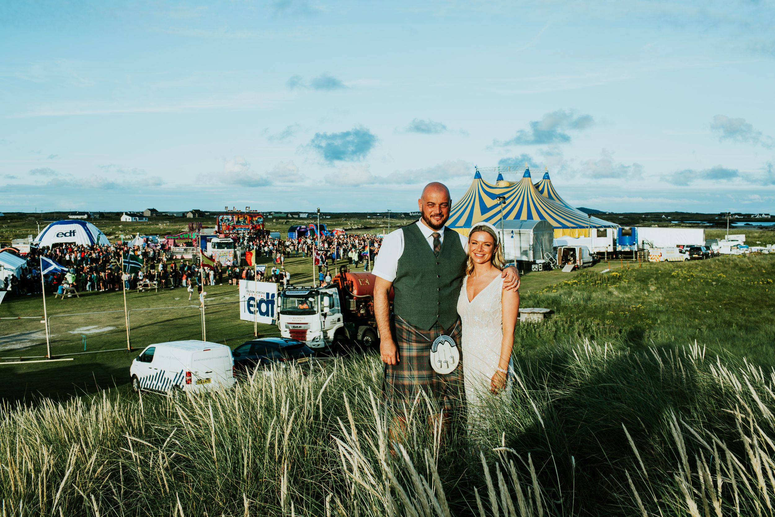 A couple stands in a coastal landscape with beach grass and tents during an outdoor event under blue skies.
