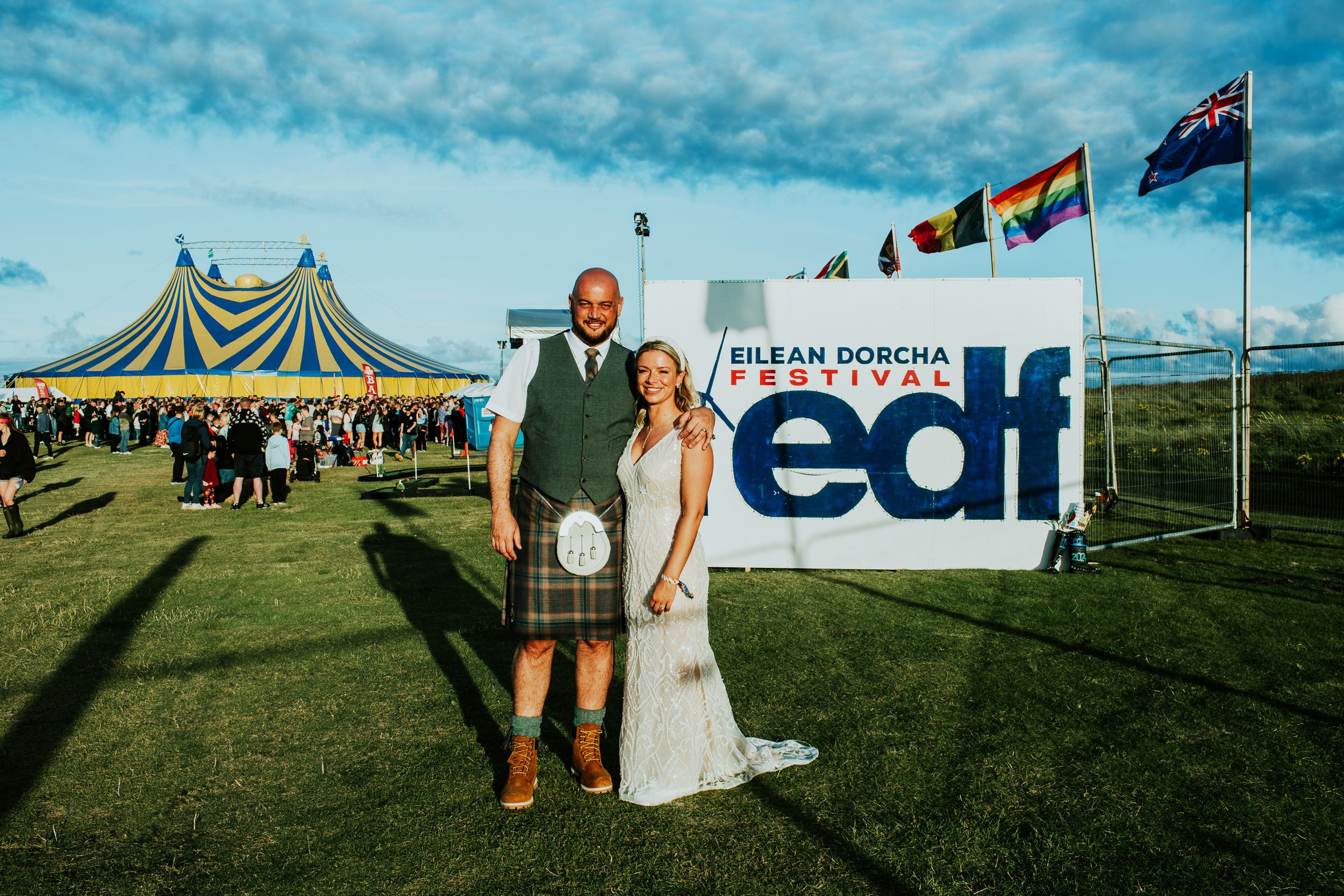 A couple poses in front of a blue and white festival banner with striped tents and flags in the background.