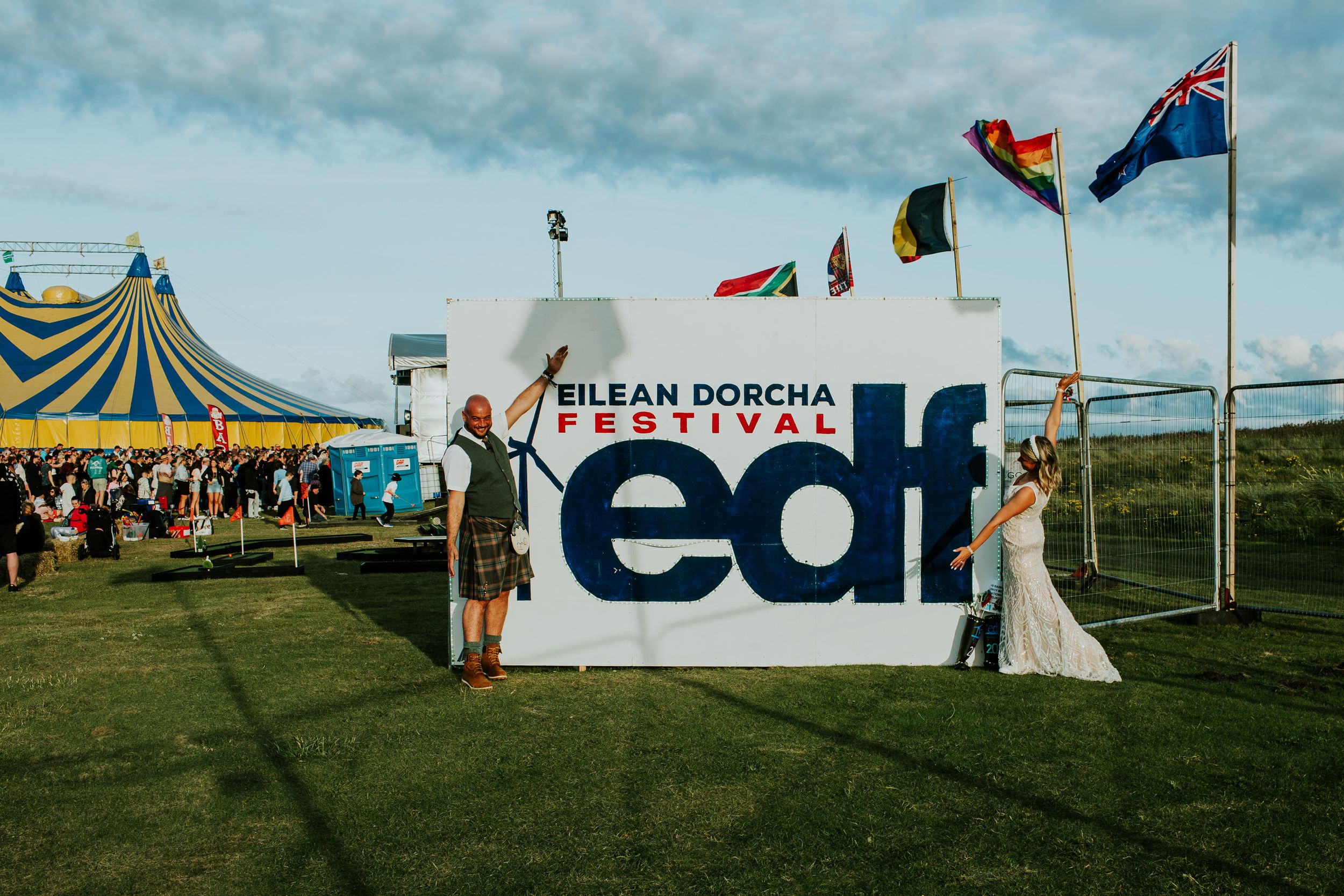 Wedding couple stands next to large 'edf' festival sign with international flags flying overhead.