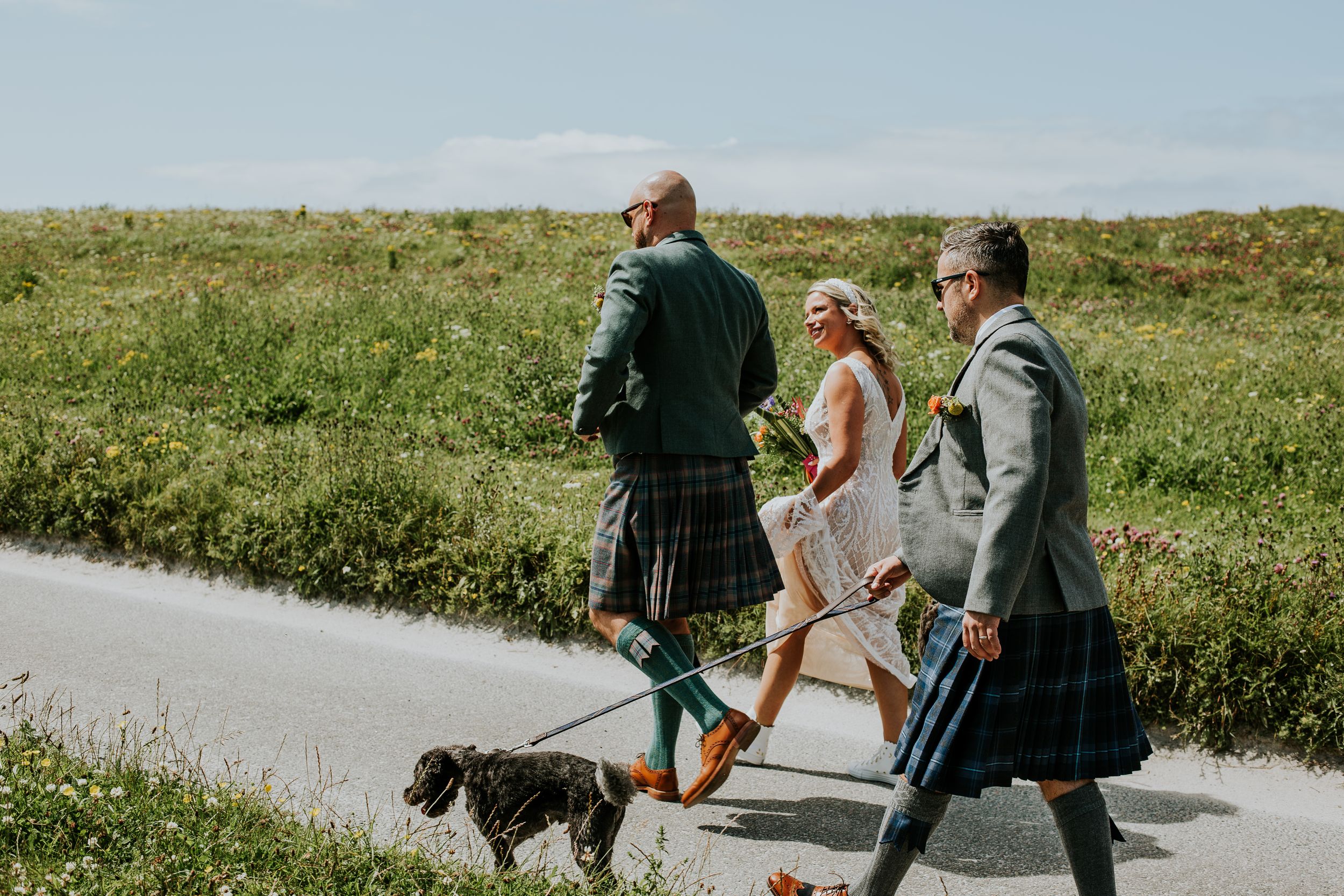 Three people in kilts walking a small dog on a paved path through green countryside on a sunny day.