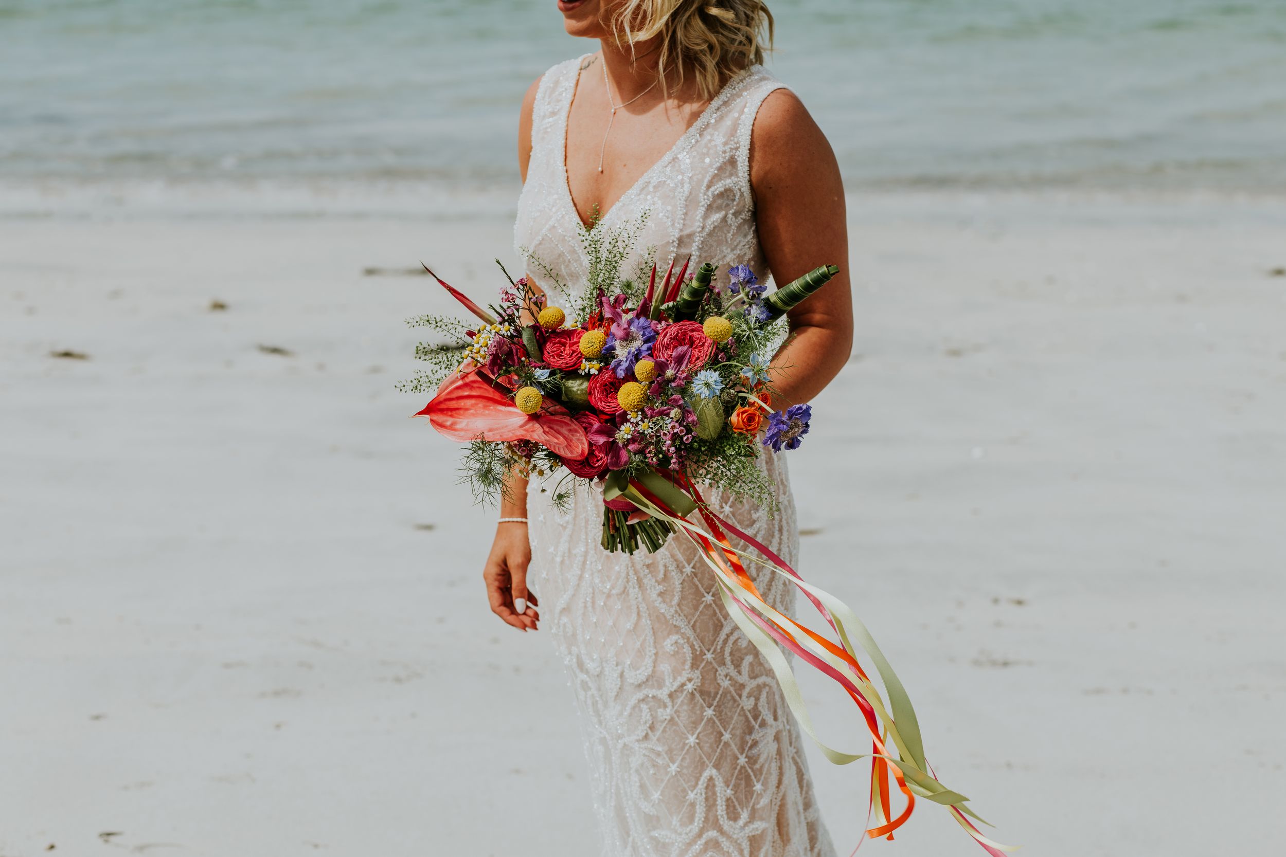 Person in lace wedding dress holds vibrant tropical bouquet with red flowers and colorful ribbons on sandy beach.