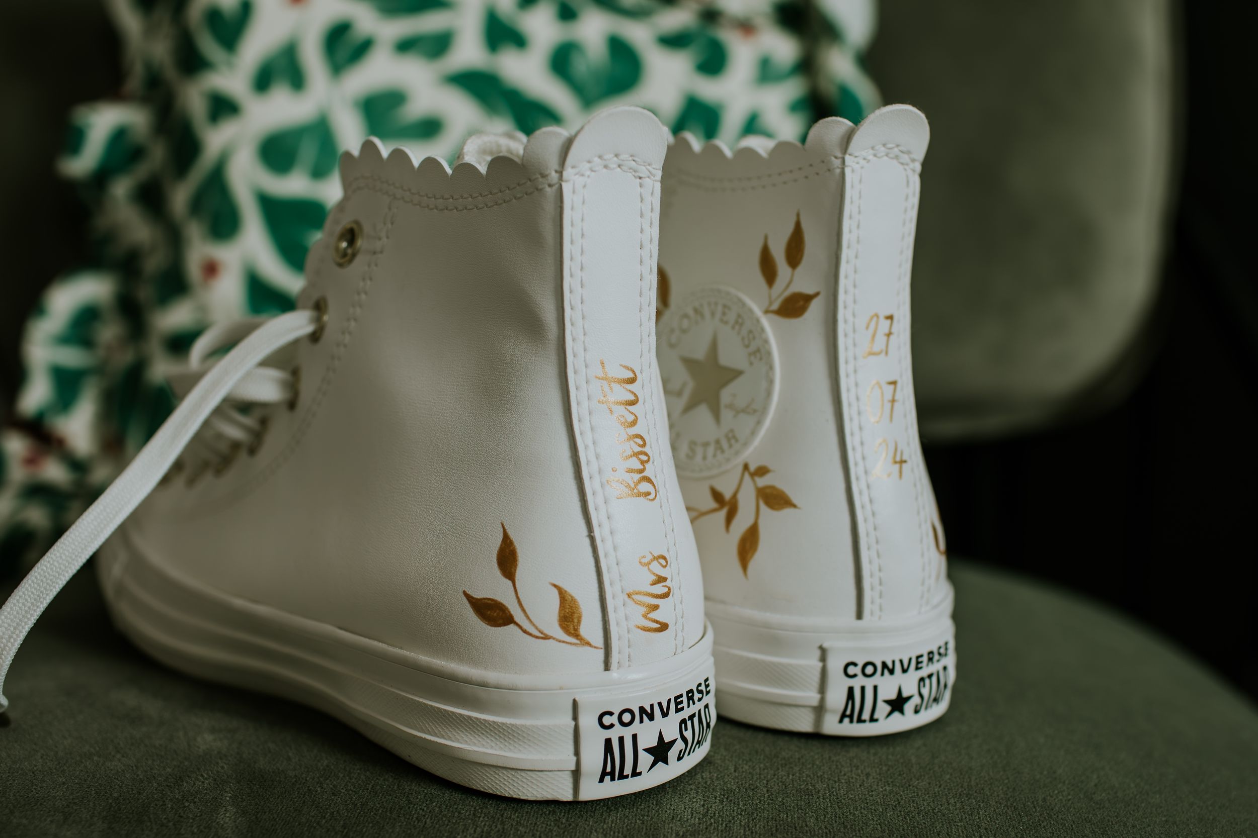 White leather high-top Converse sneaker with delicate gold floral design details sits against green background.