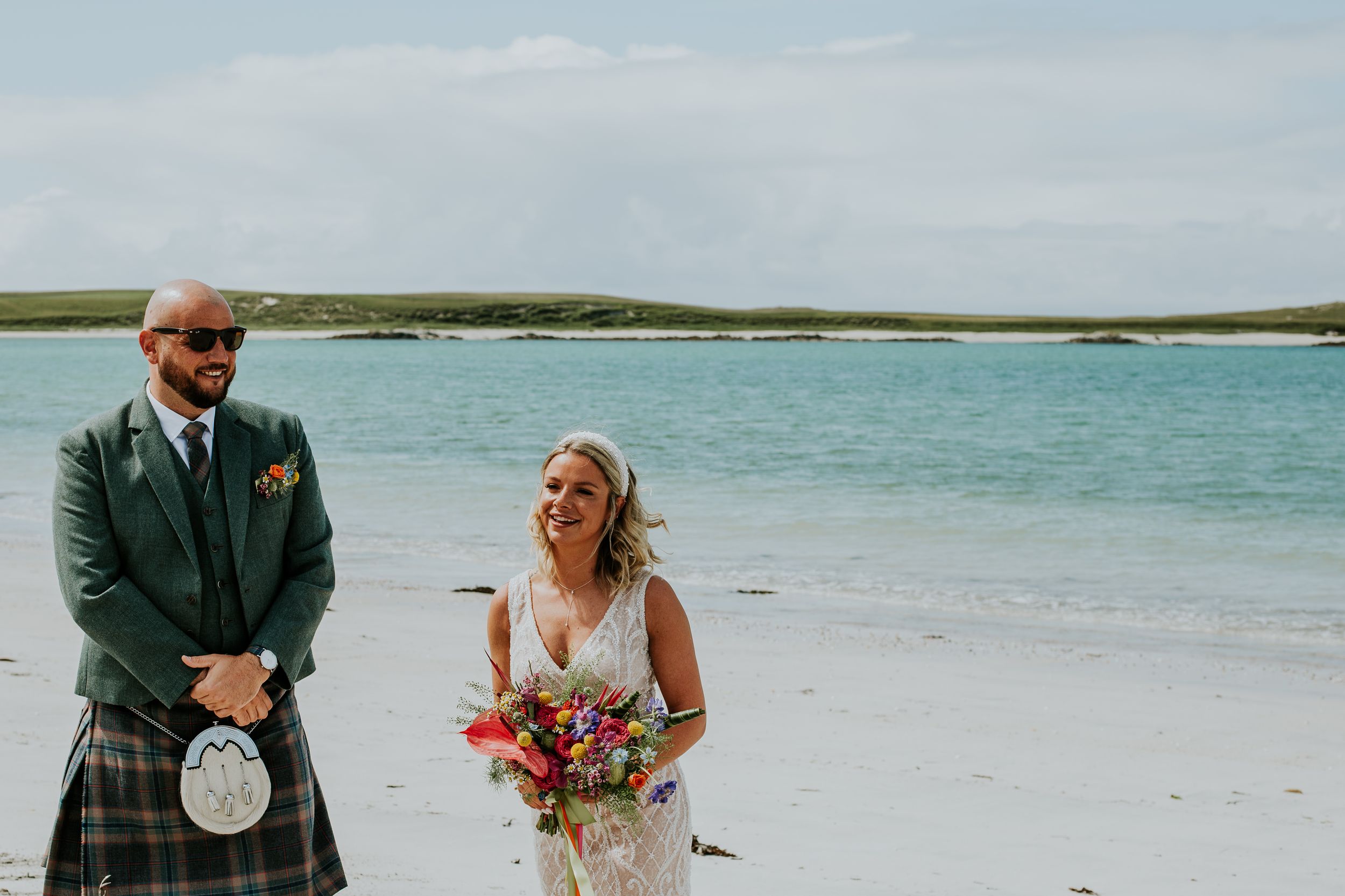 Two people on a scenic beach with turquoise water, with one wearing a traditional Scottish kilt and sporran.