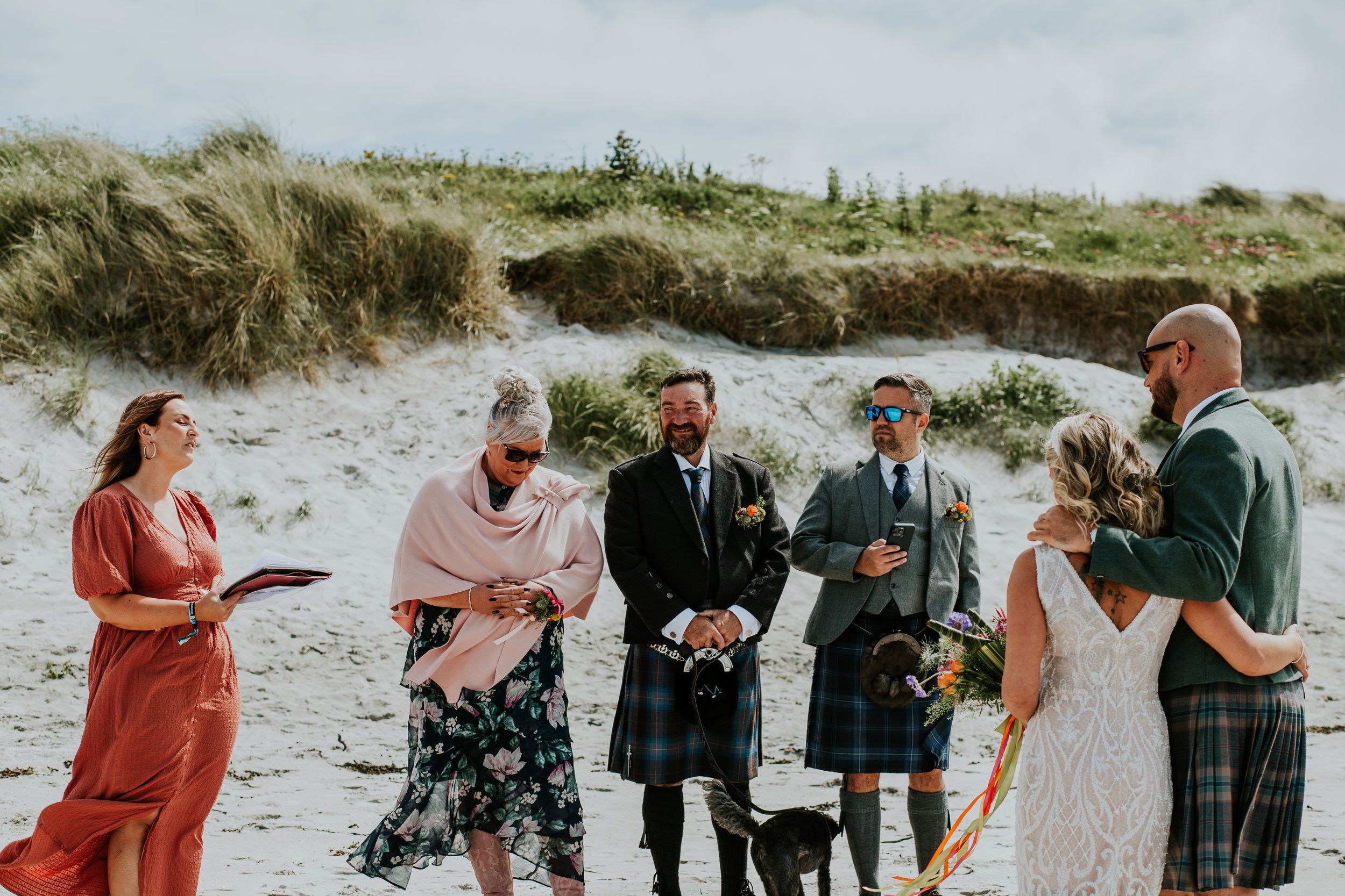 A beach wedding ceremony with guests wearing traditional Scottish kilts on a sandy shore.