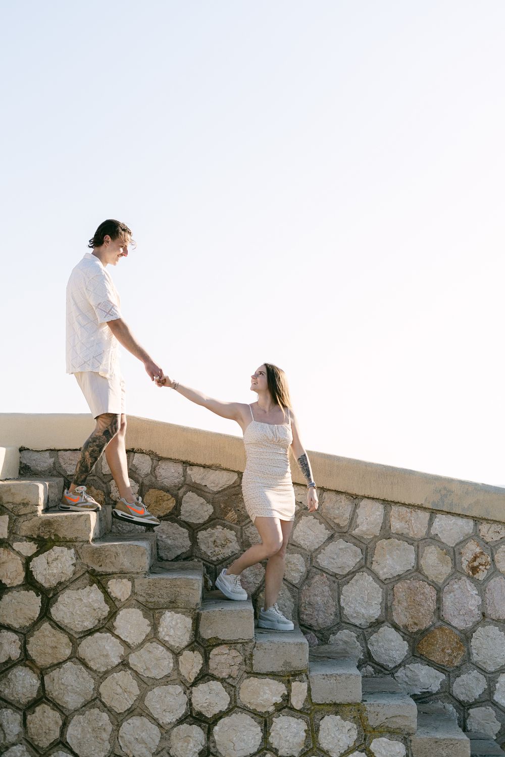 Two people in white outfits hold hands while climbing stone steps against a bright sky backdrop.