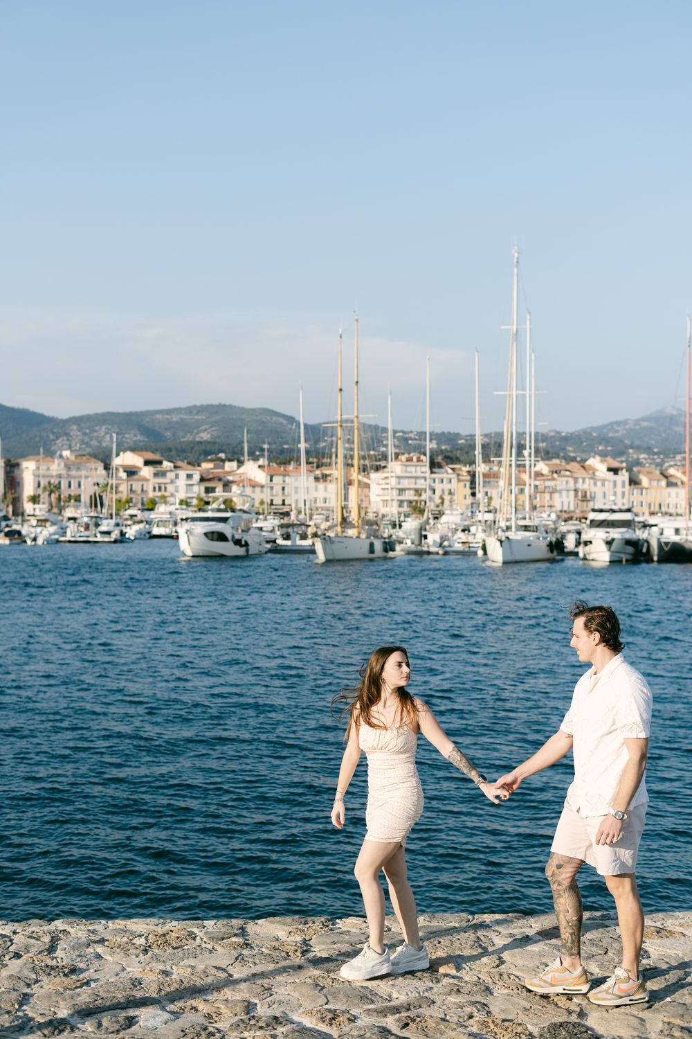Couple walking hand in hand along Mediterranean harbor with sailboats and coastal town buildings in background.