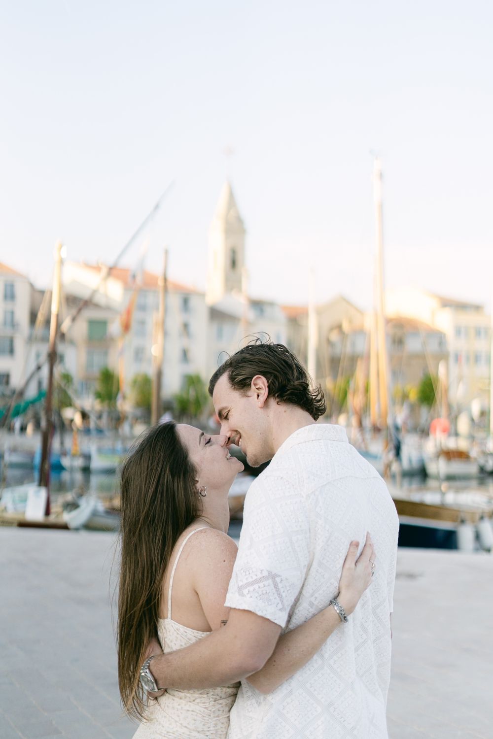 Romantic couple embracing at a Mediterranean marina with boats and old town buildings in the background at sunset.