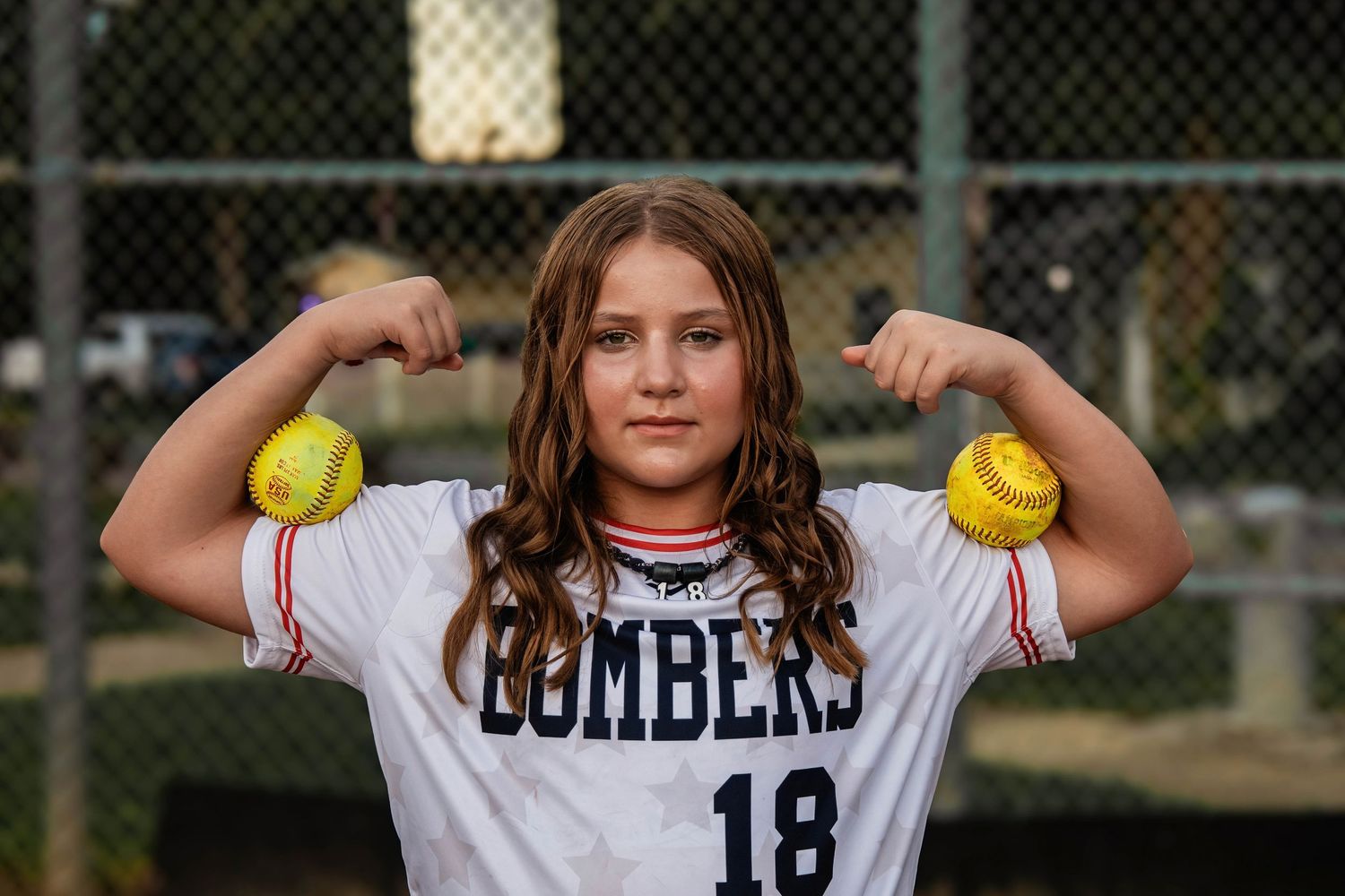Softball player wearing Bombers jersey #18 flexes while holding yellow softballs at baseball field.