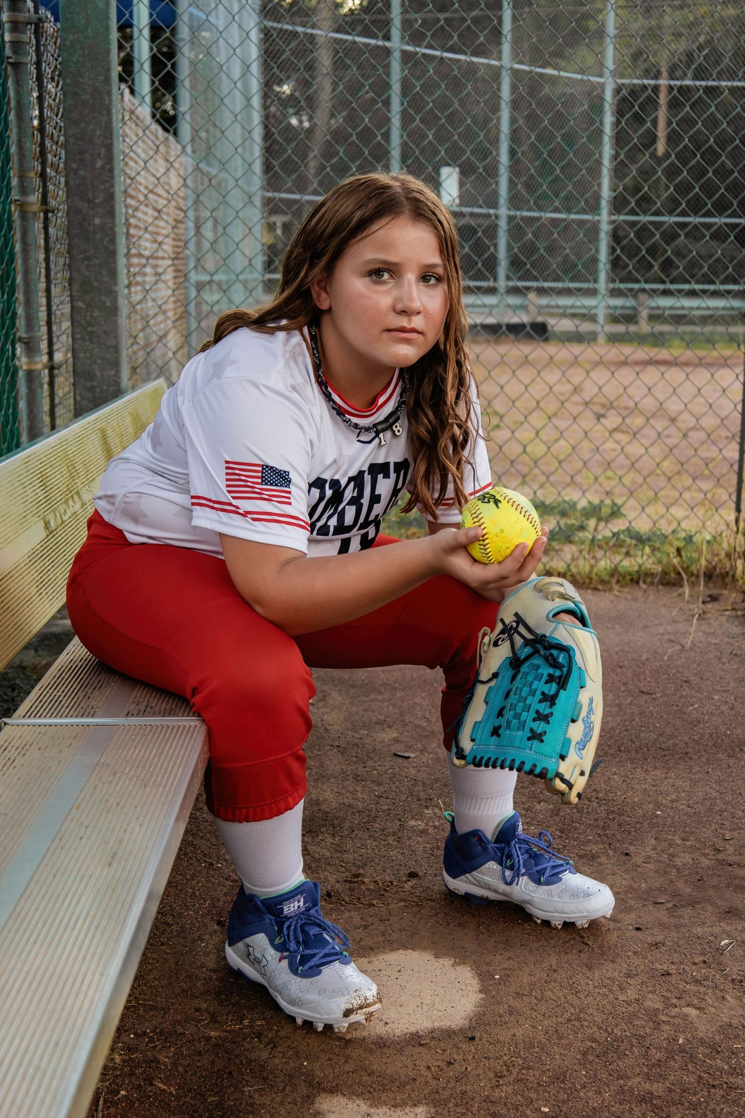 Softball player in Bombers uniform sits on bench holding yellow ball at field.