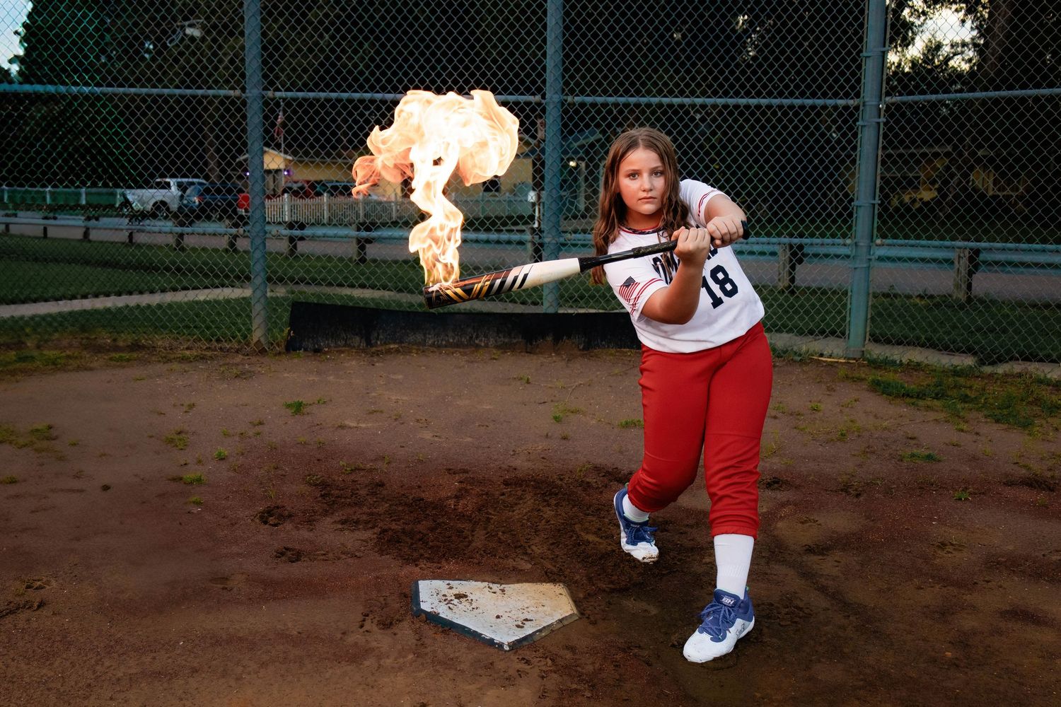 Softball player in uniform poses with flaming bat on baseball diamond at dusk.