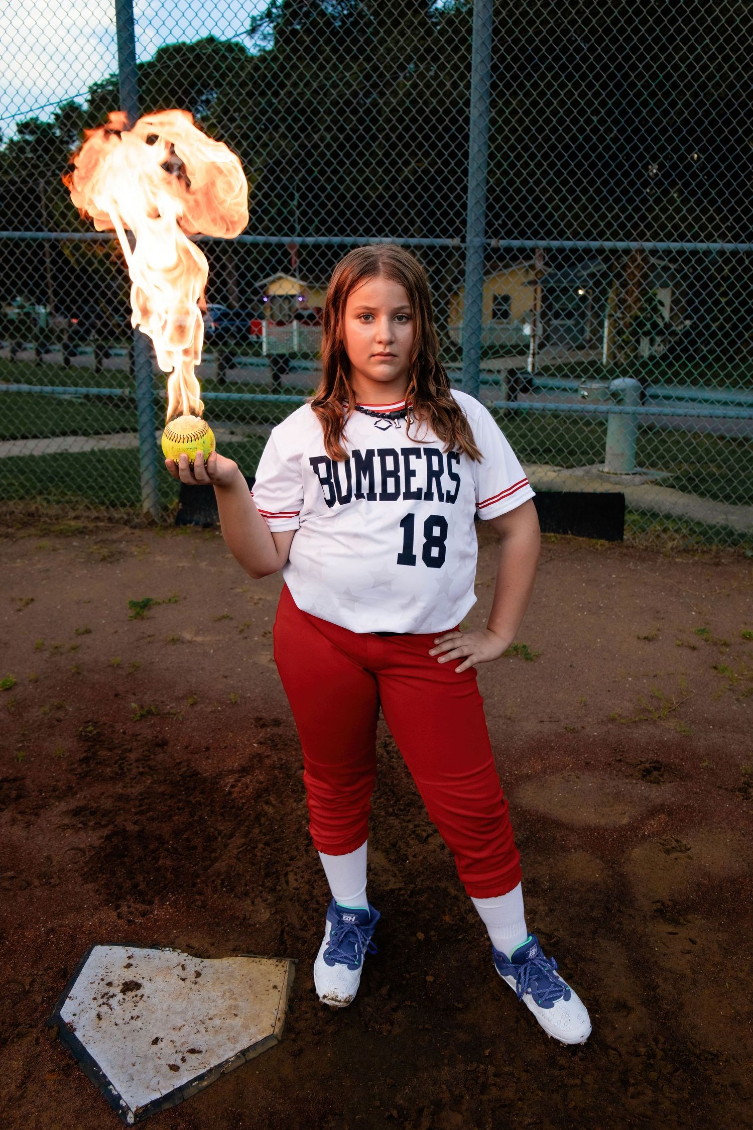 Softball player in Bombers uniform holds yellow ball engulfed in flames on baseball field.