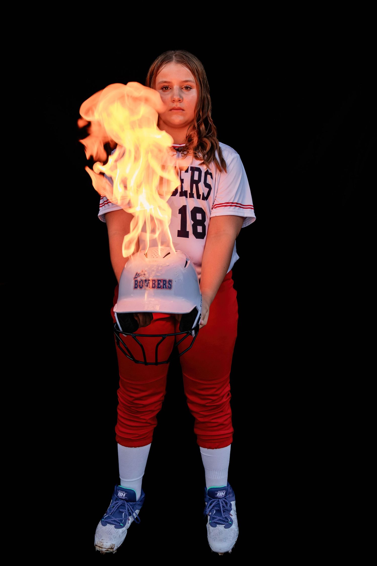 Softball player poses with flaming equipment against dark background in dramatic portrait.